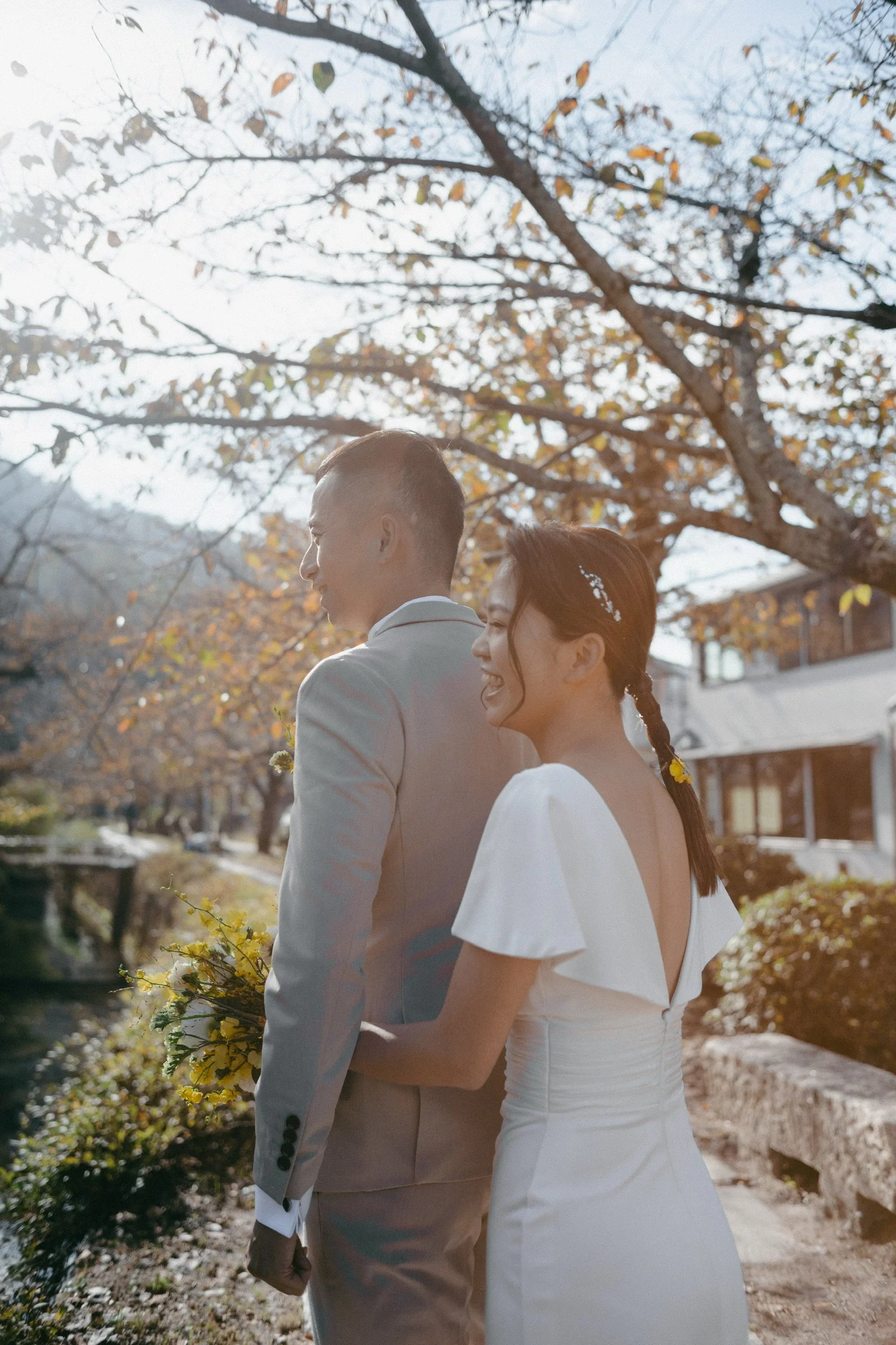 A couple dressed in wedding attire walking outdoors under leafless trees during autumn, with a small river or creek nearby and sunlight filtering through the branches.