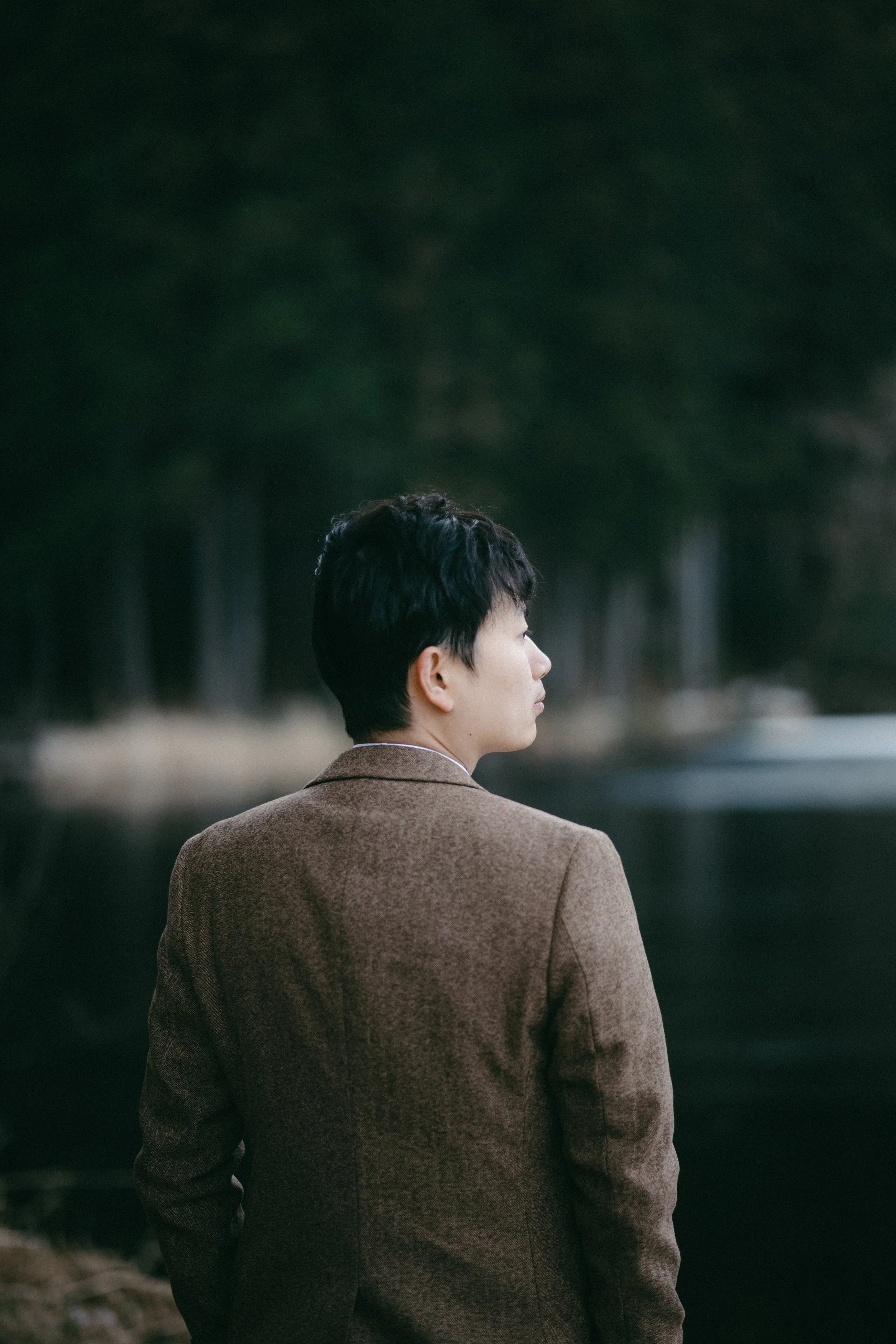 Side view of a woman in a brown blazer standing near water with trees in the background.
