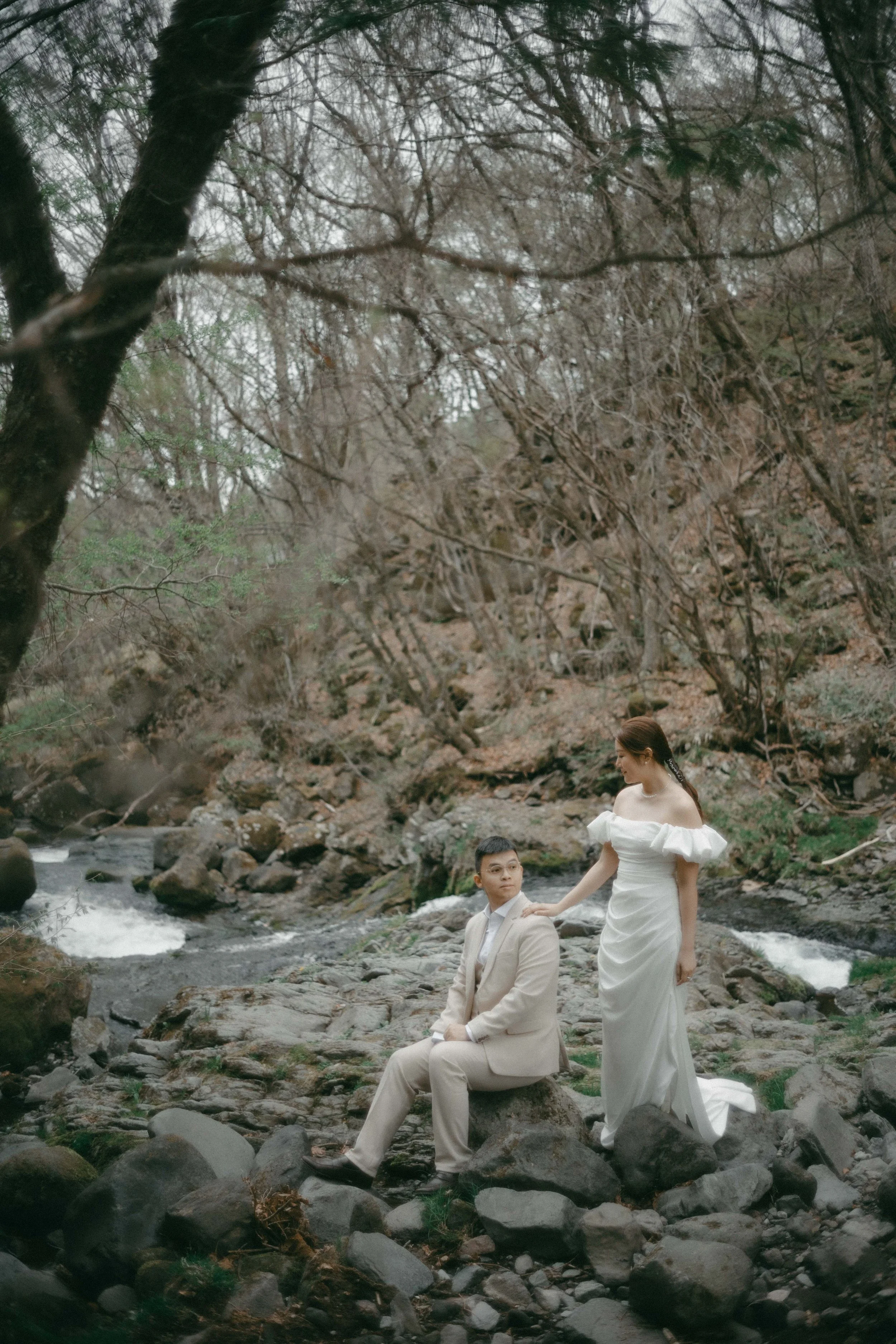 A couple in wedding attire in a mountainous, forested area by a stream. The woman is standing, wearing a white off-shoulder wedding dress with puffed sleeves, resting her hand on the seated man's shoulder. The man is dressed in a beige suit, seated o