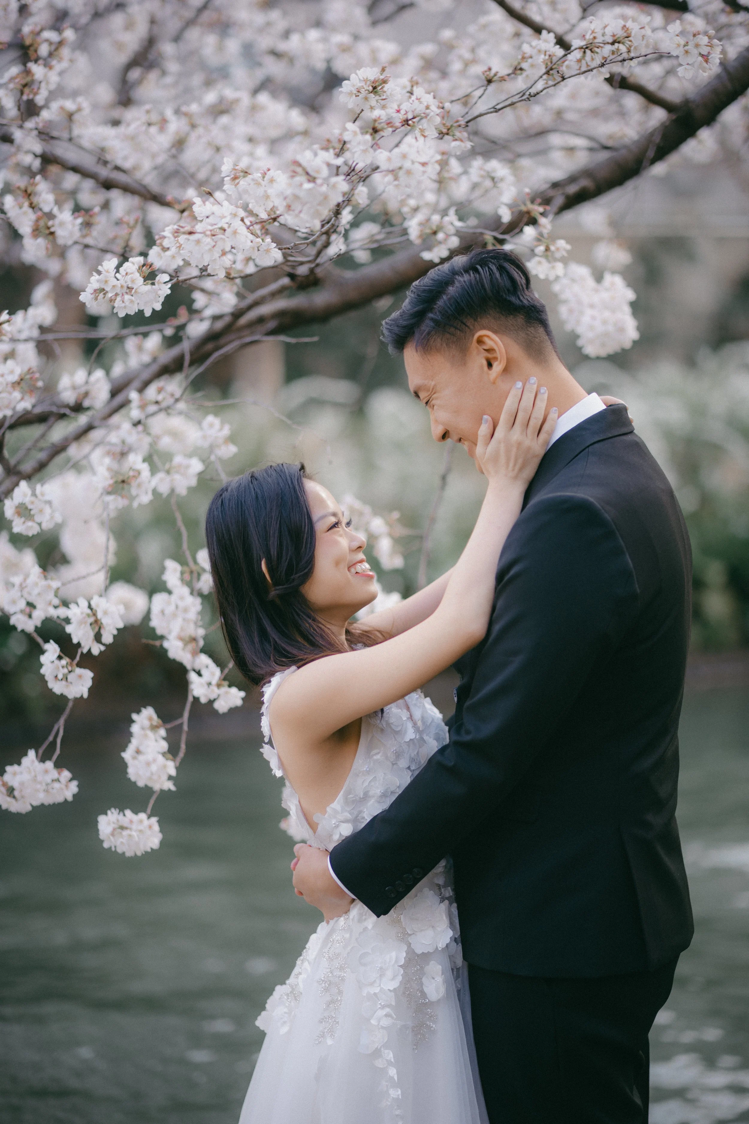 A couple dressed in wedding attire, embracing and smiling at each other under blooming cherry blossom trees near a body of water.