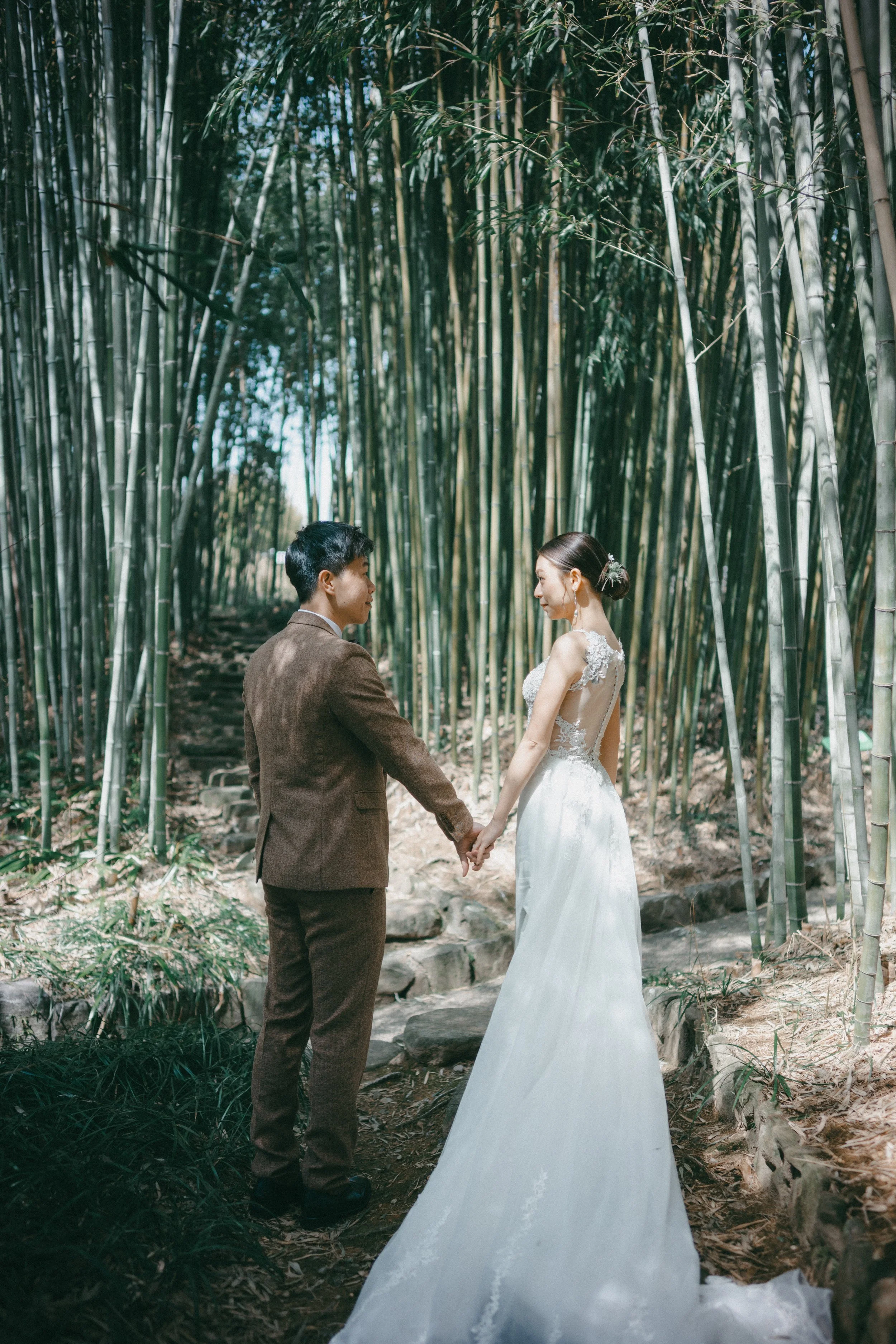 A bride and groom holding hands in a bamboo forest, with the bride in a white wedding dress and the groom in a brown suit.