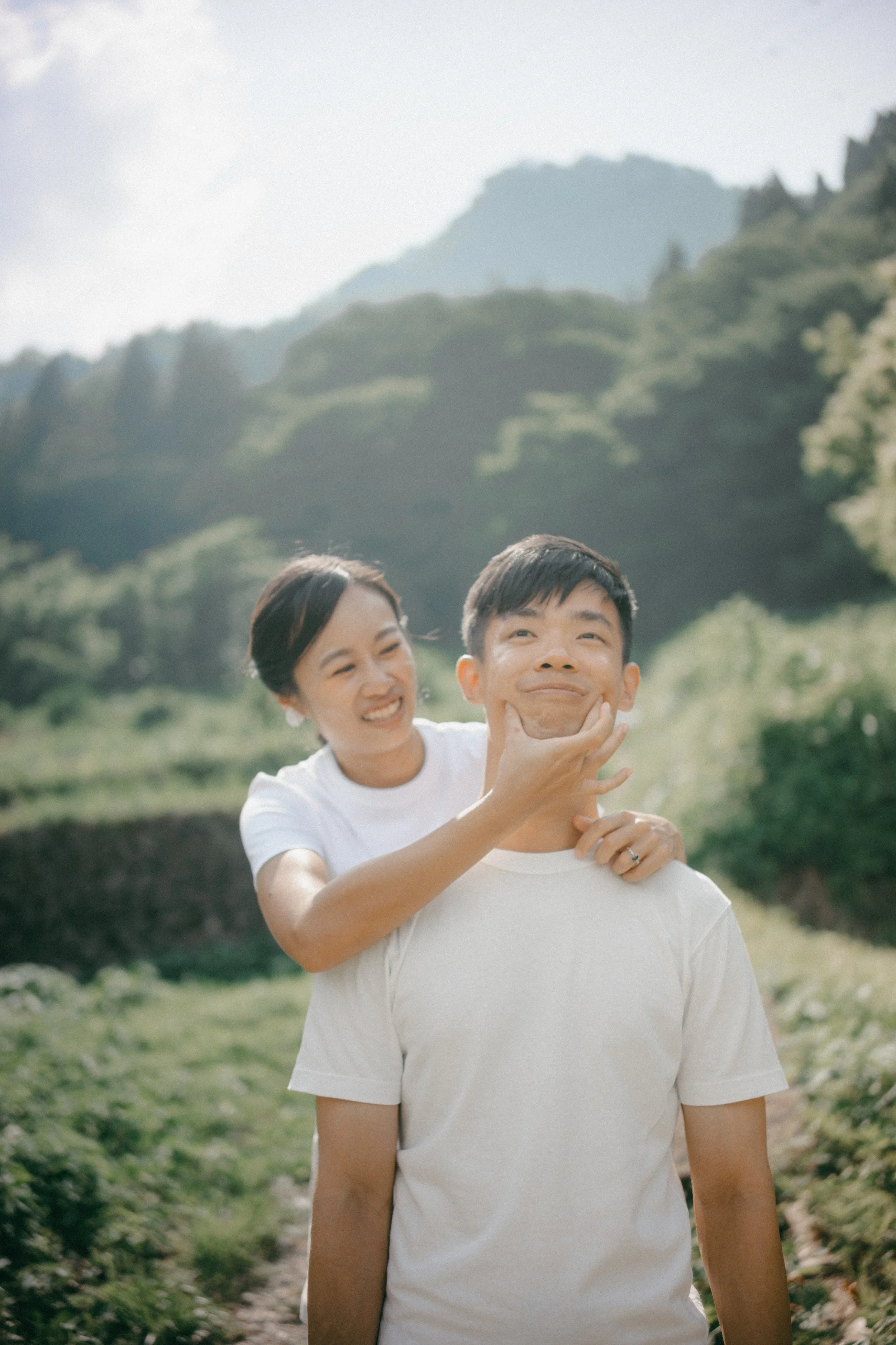 A woman playful pinches a man's chin from behind in a scenic outdoor setting with mountains and greenery in the background.
