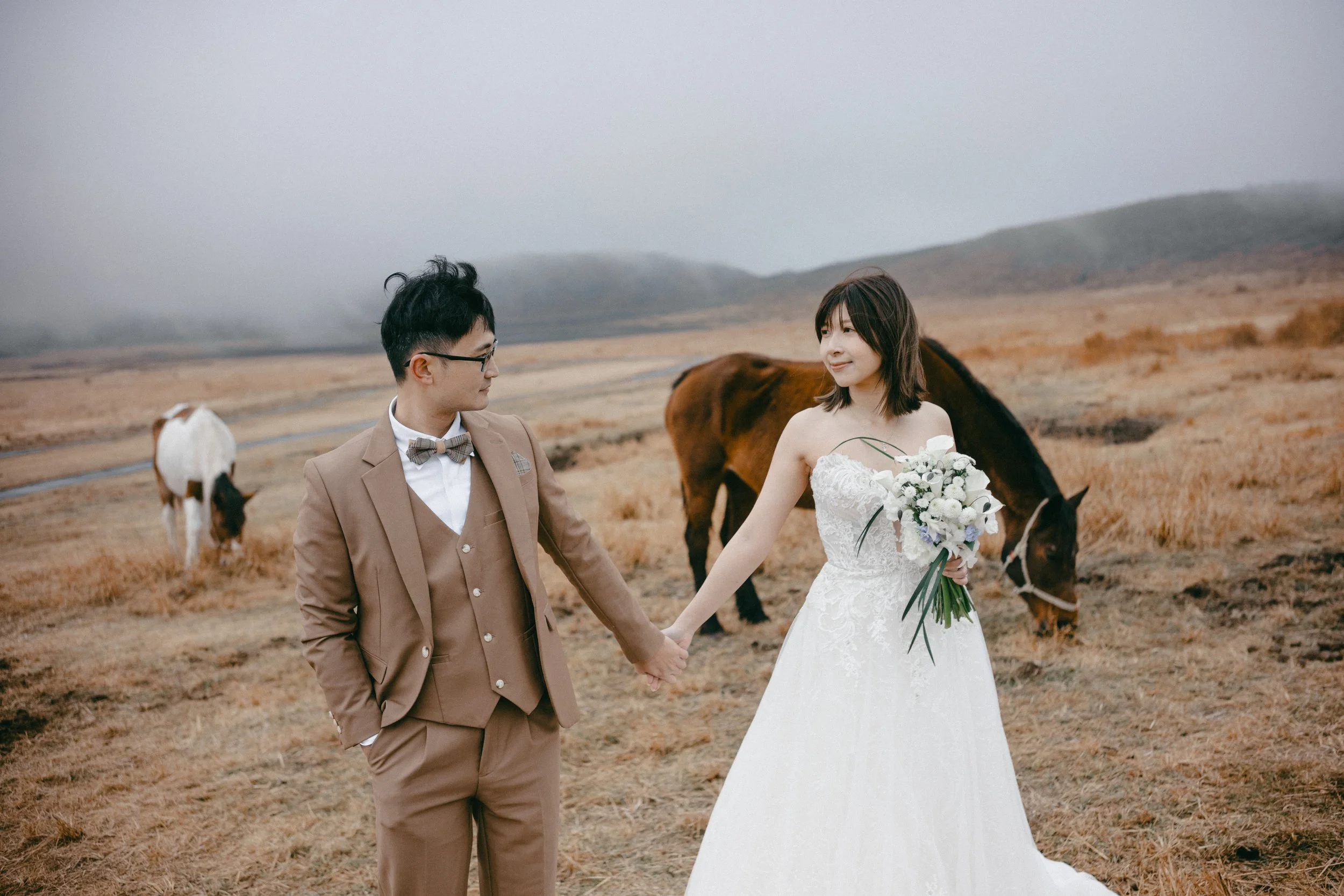 A bride and groom holding hands in a field with horses, with misty hills in the background.