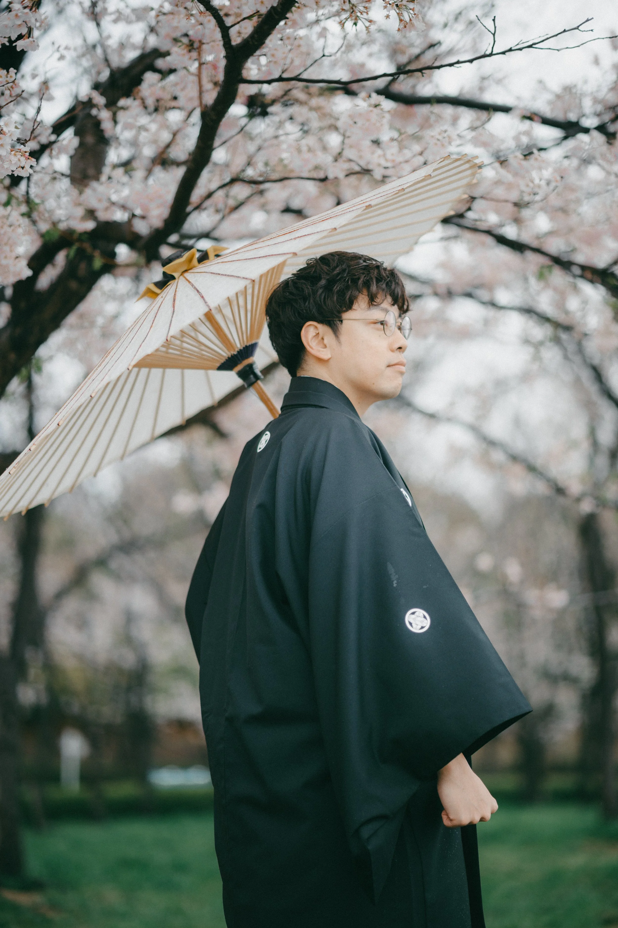A man wearing traditional Japanese clothing holding an umbrella stands near cherry blossom trees.