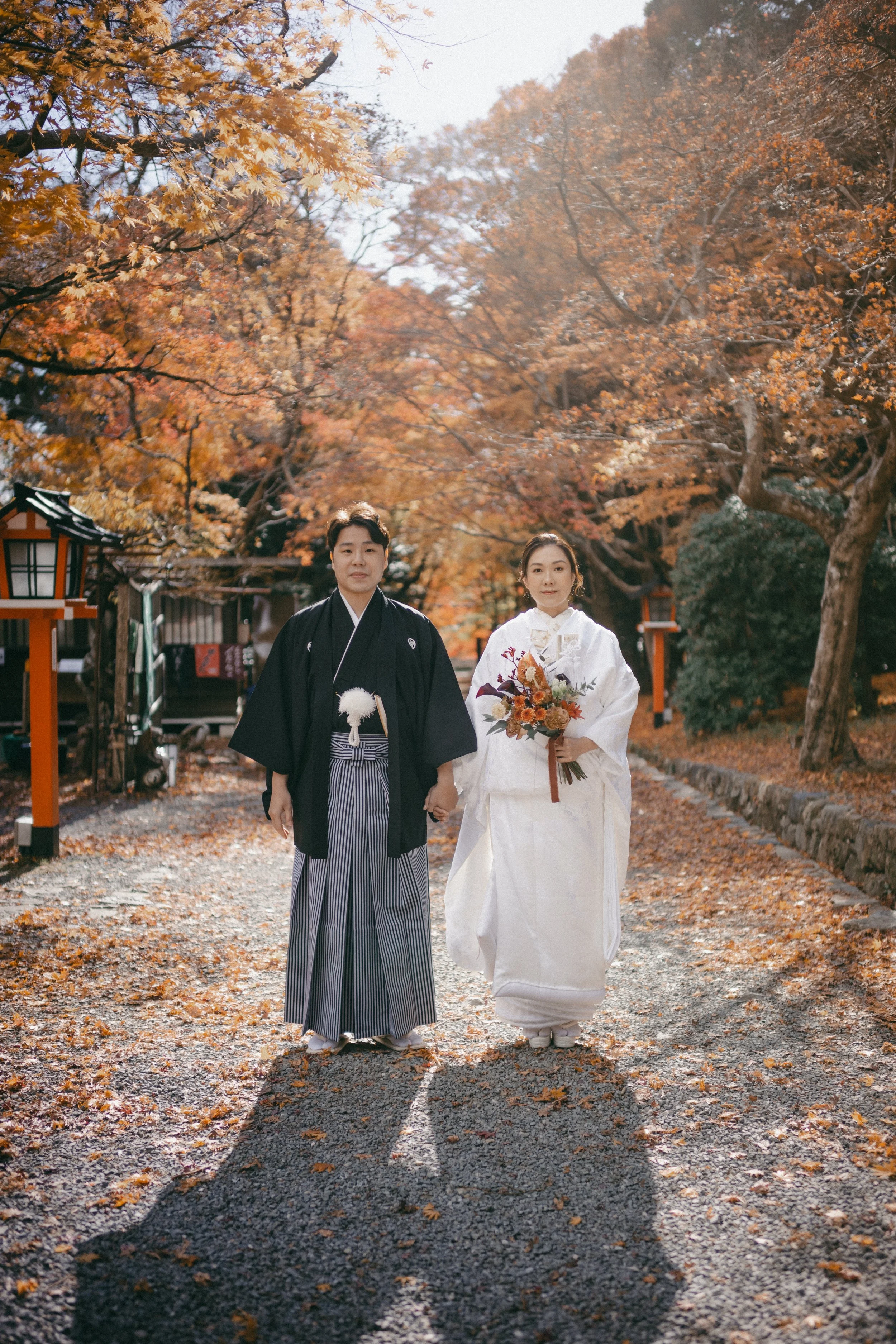 A bride and groom walking together in traditional Japanese wedding attire, holding hands, surrounded by autumn trees and a gravel path.