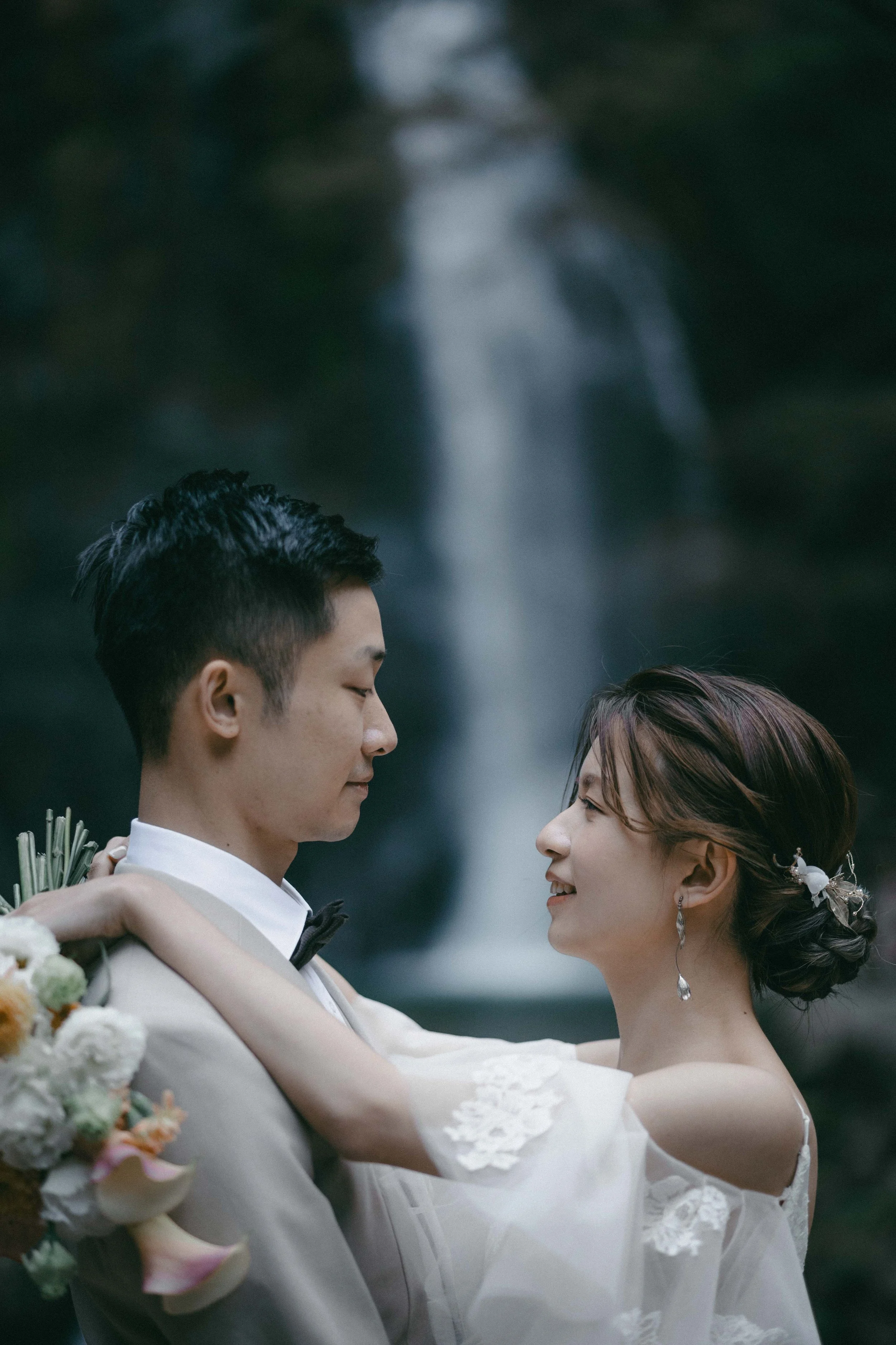 A bride and groom embracing outdoors with a waterfall in the background.