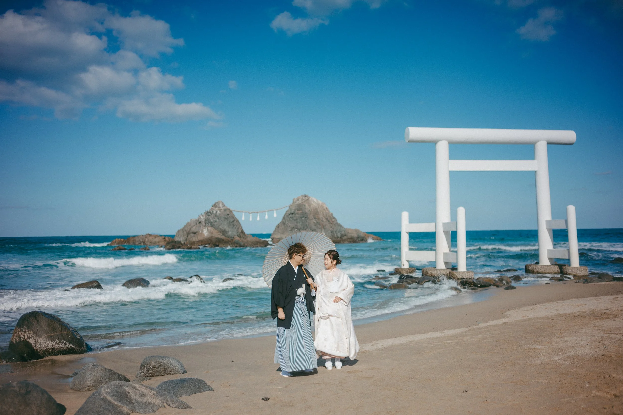 Two people dressed in traditional Japanese wedding attire standing on a beach near the ocean with rocks and a large white torii gate in the background.