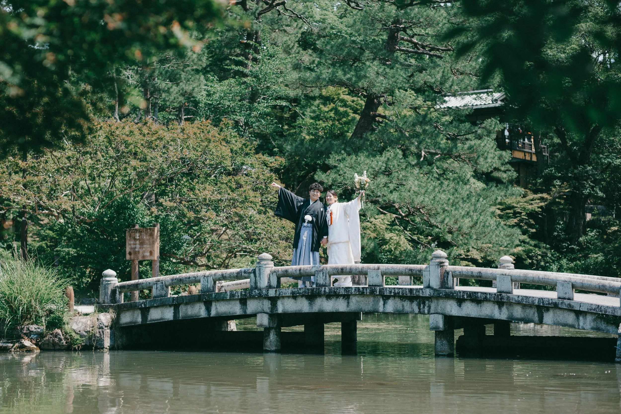 A newlywed couple in traditional Japanese wedding attire standing on a stone bridge over a pond, waving and smiling, surrounded by lush green trees in a serene outdoor setting.