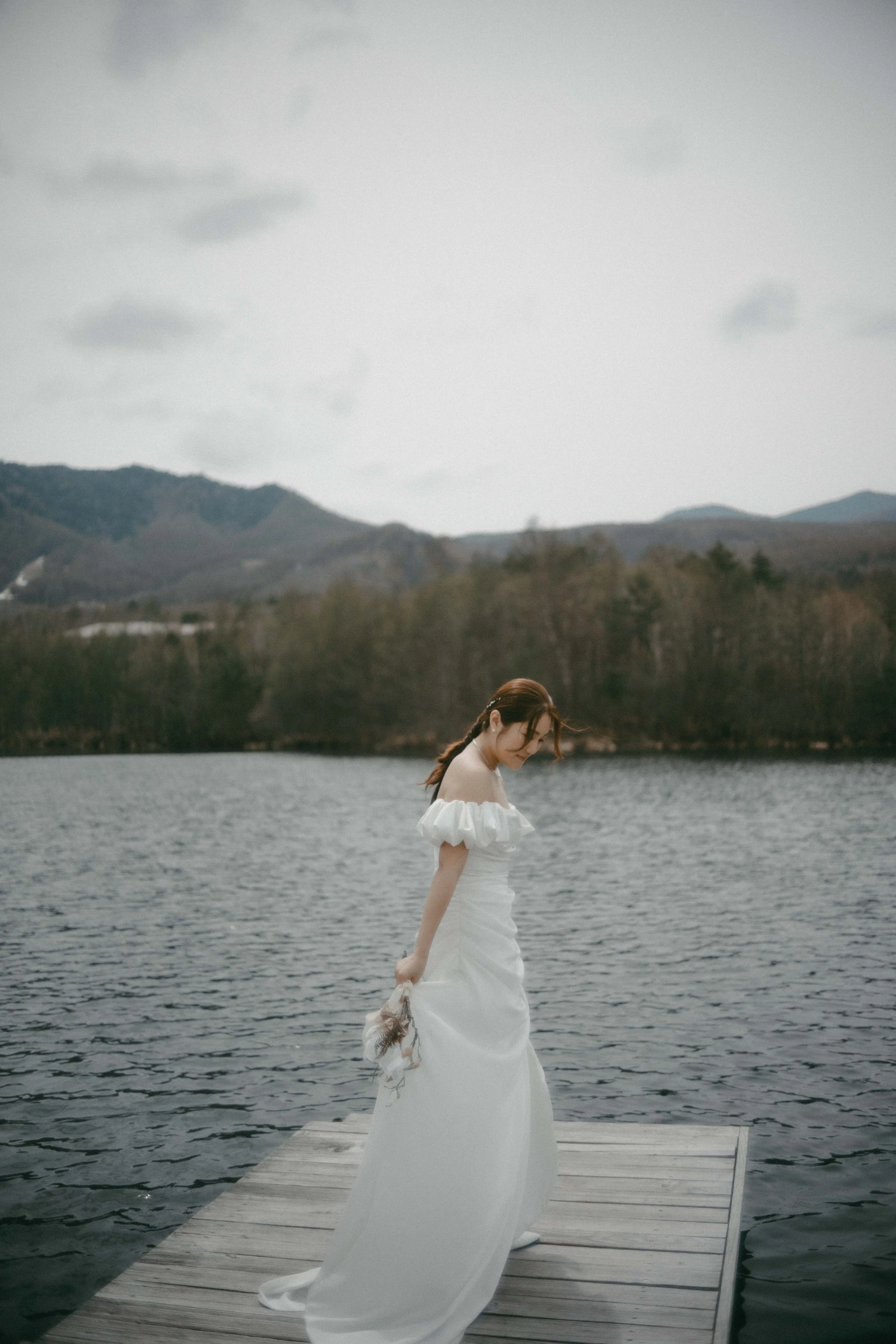 A woman in a white off-shoulder wedding dress holding a bouquet, standing on a wooden dock by a lake, with mountains and cloudy sky in the background.