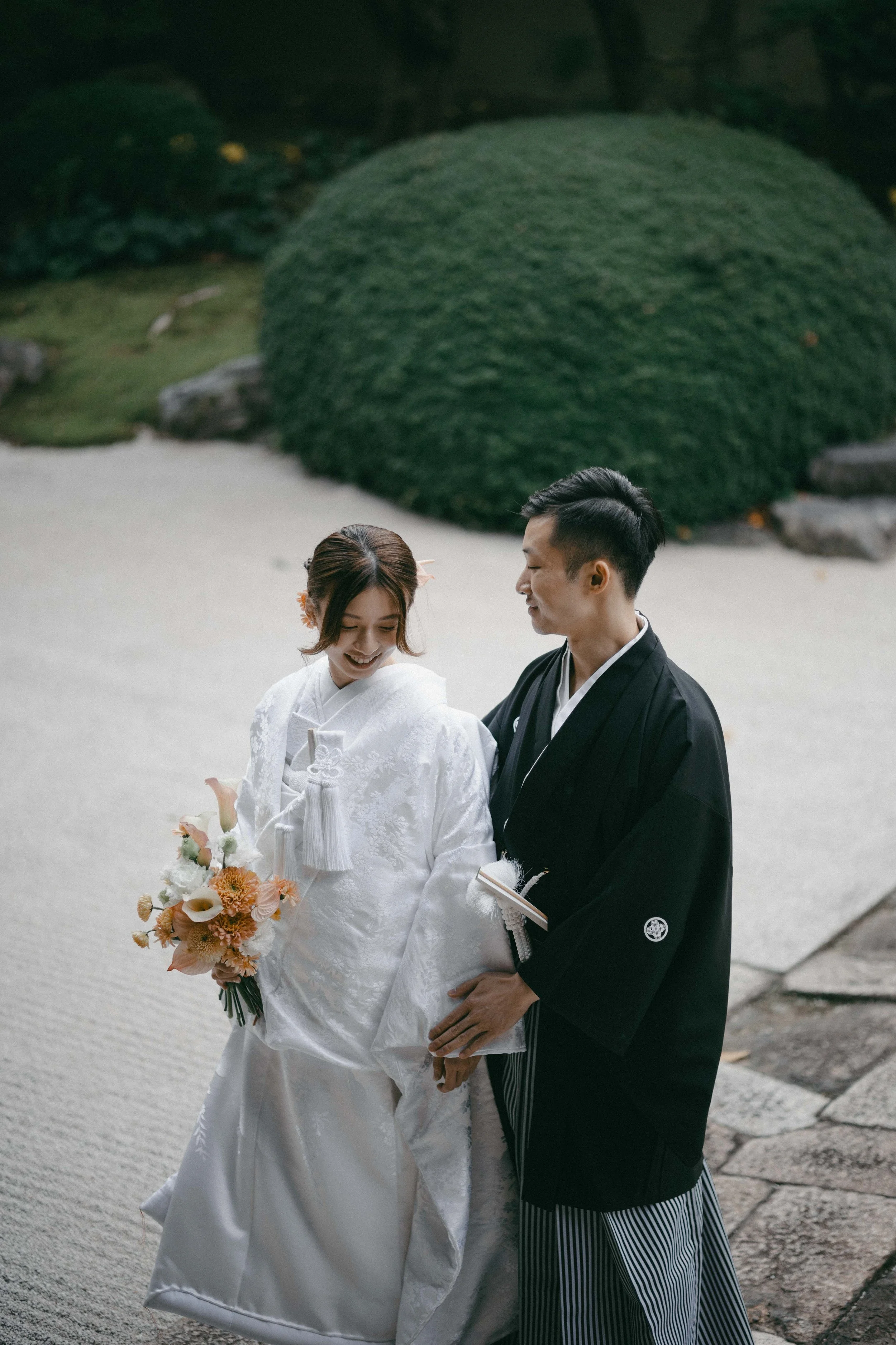 A bride and groom in traditional Japanese wedding attire, standing outdoors on a stone pathway with a large green bush in the background. The bride is holding a bouquet of flowers and smiling, while the groom is looking at her.