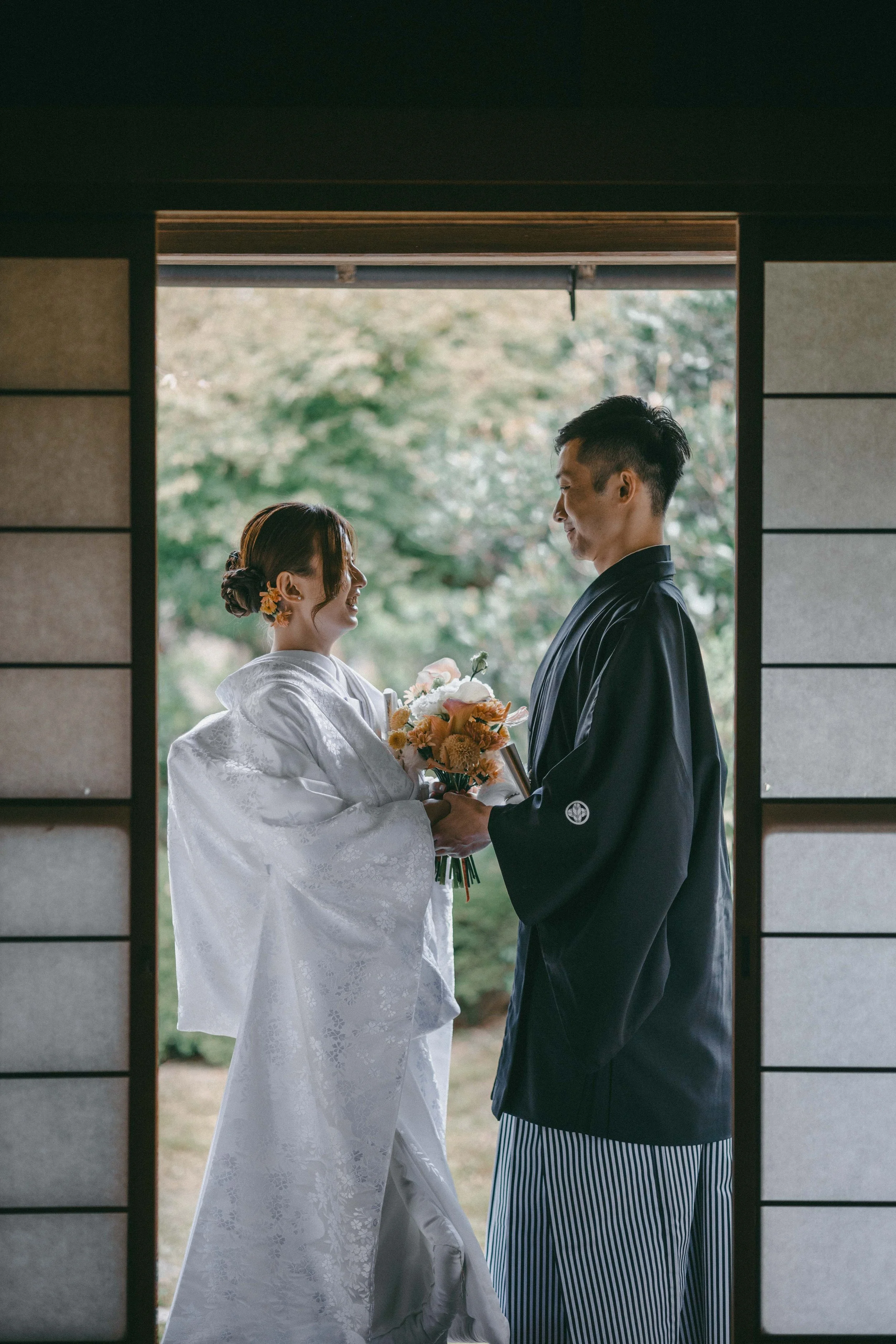 A couple in traditional Japanese wedding attire standing inside a house, holding a bouquet of flowers, and smiling at each other during a wedding ceremony.