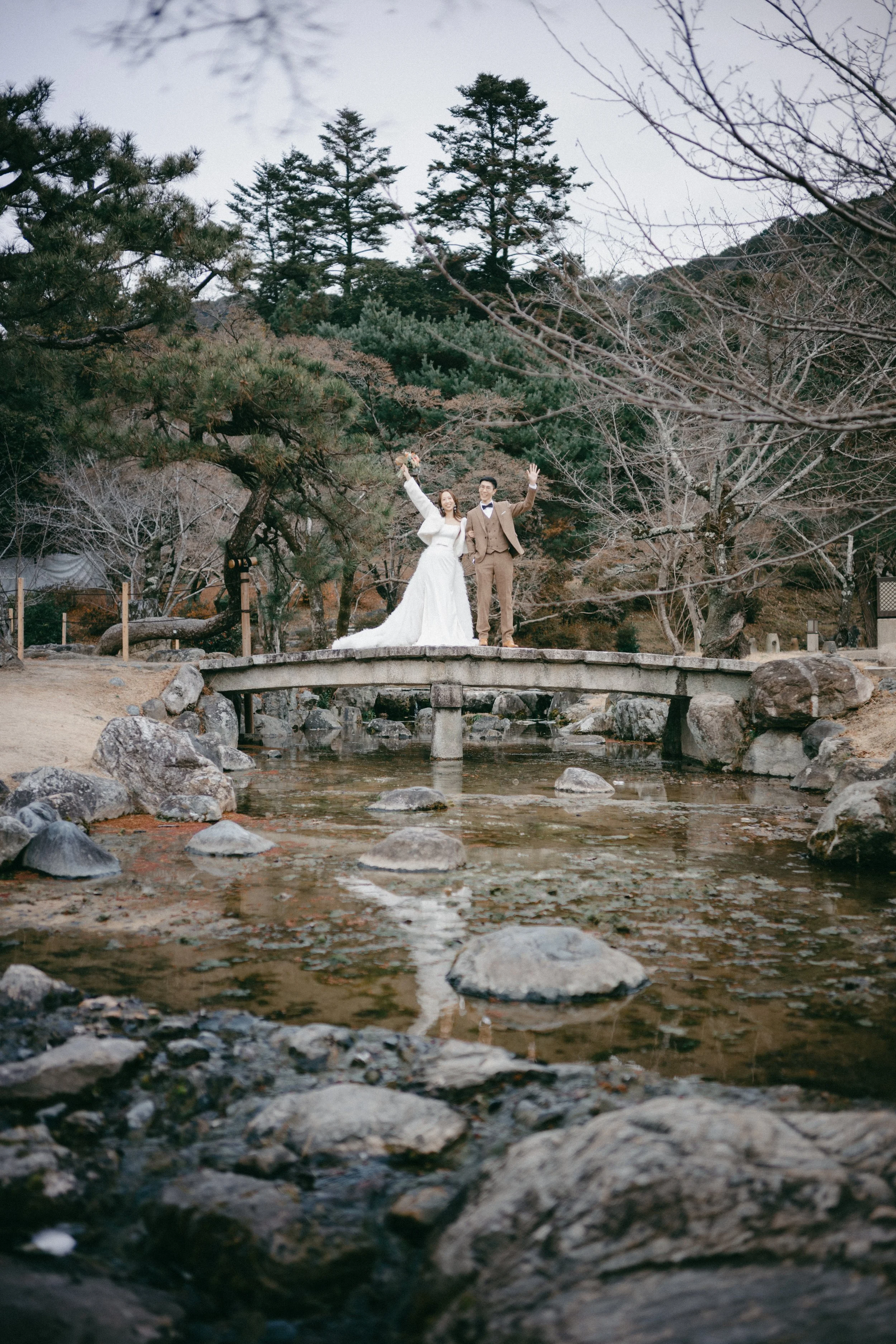 A bride and groom standing on a small stone bridge over a creek in a natural park setting with trees and rocks, celebrating their wedding.