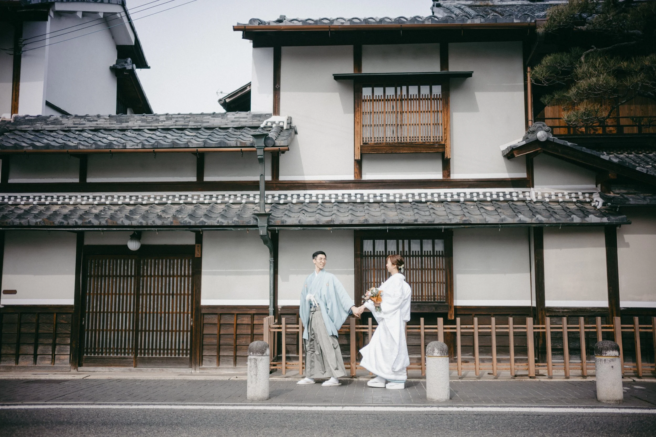 A bride and groom dressed in traditional Japanese wedding attire walking hand in hand on a street in front of a traditional Japanese building.