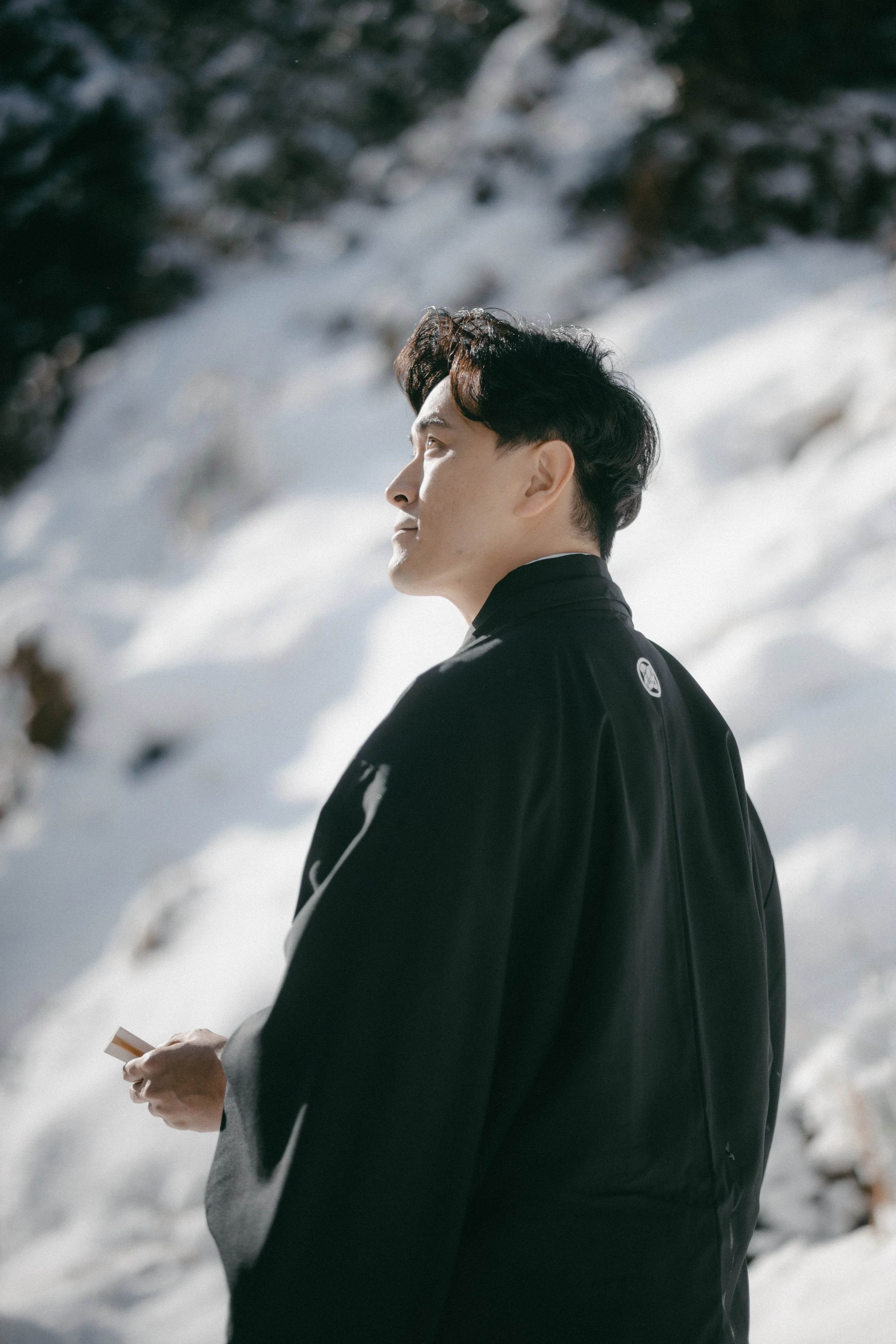 A man standing outdoors in a snowy landscape, dressed in a black jacket, holding an object in his hand, and looking to the left with mountains and trees in the background.