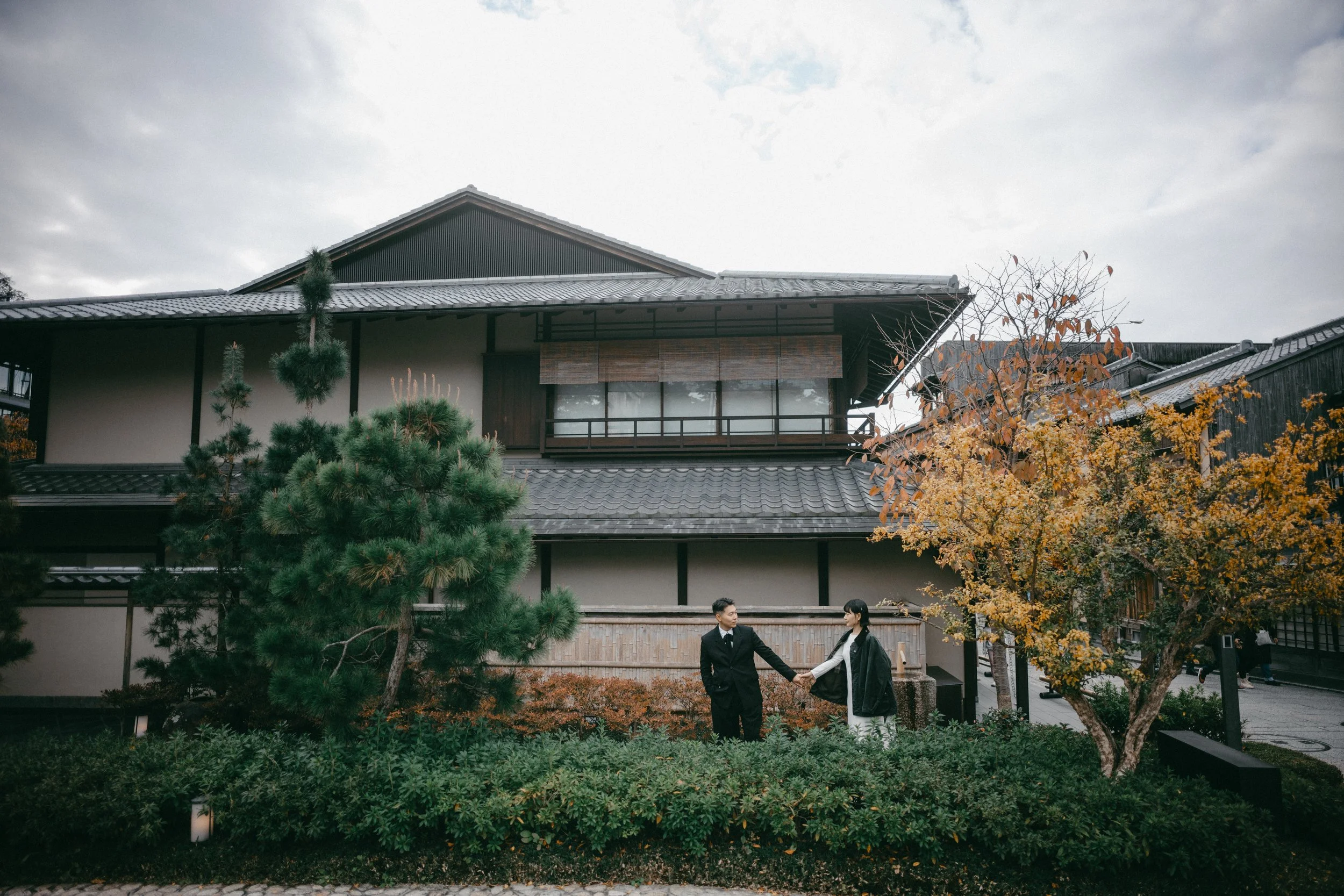 A couple dressed in formal attire holding hands in front of a traditional Japanese building with trees and greenery around them.