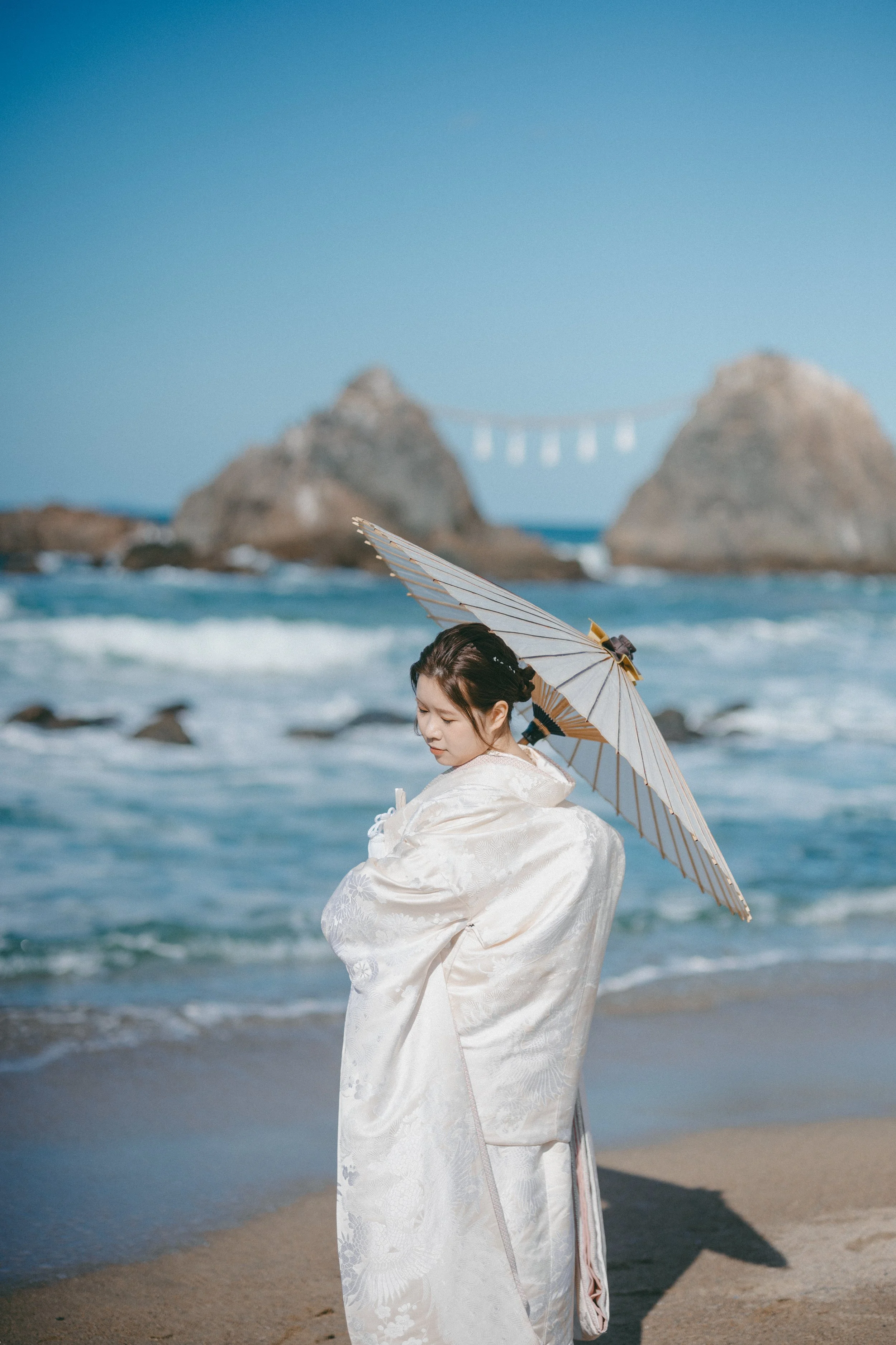 A woman dressed in traditional Japanese attire holding a paper parasol on a beach with waves, large rocks, and a distant bridge in the background.
