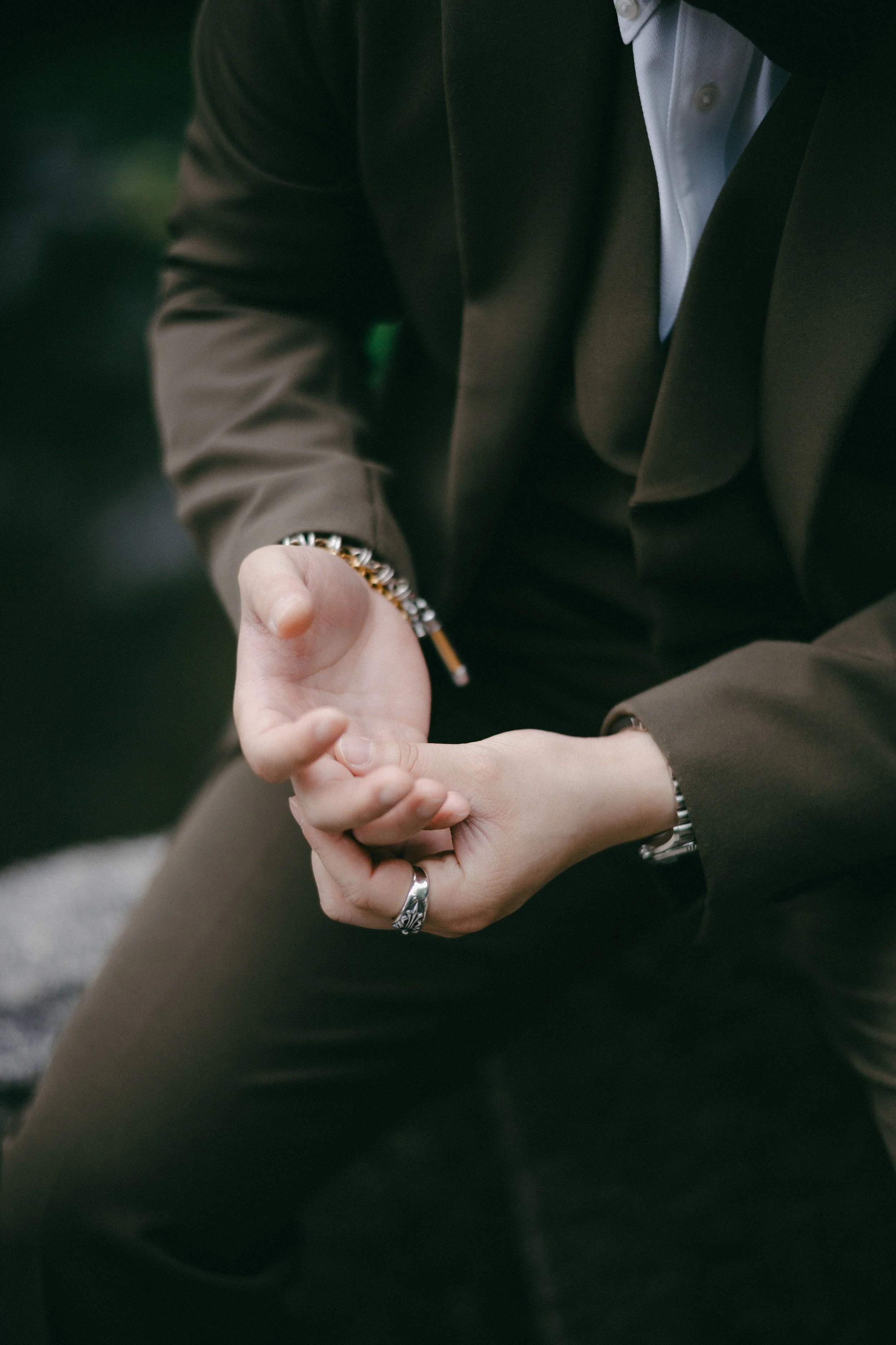 A person wearing a brown suit and white shirt, sitting outdoors, with their hands clasped together and near their knee, wearing a silver ring and bracelet.
