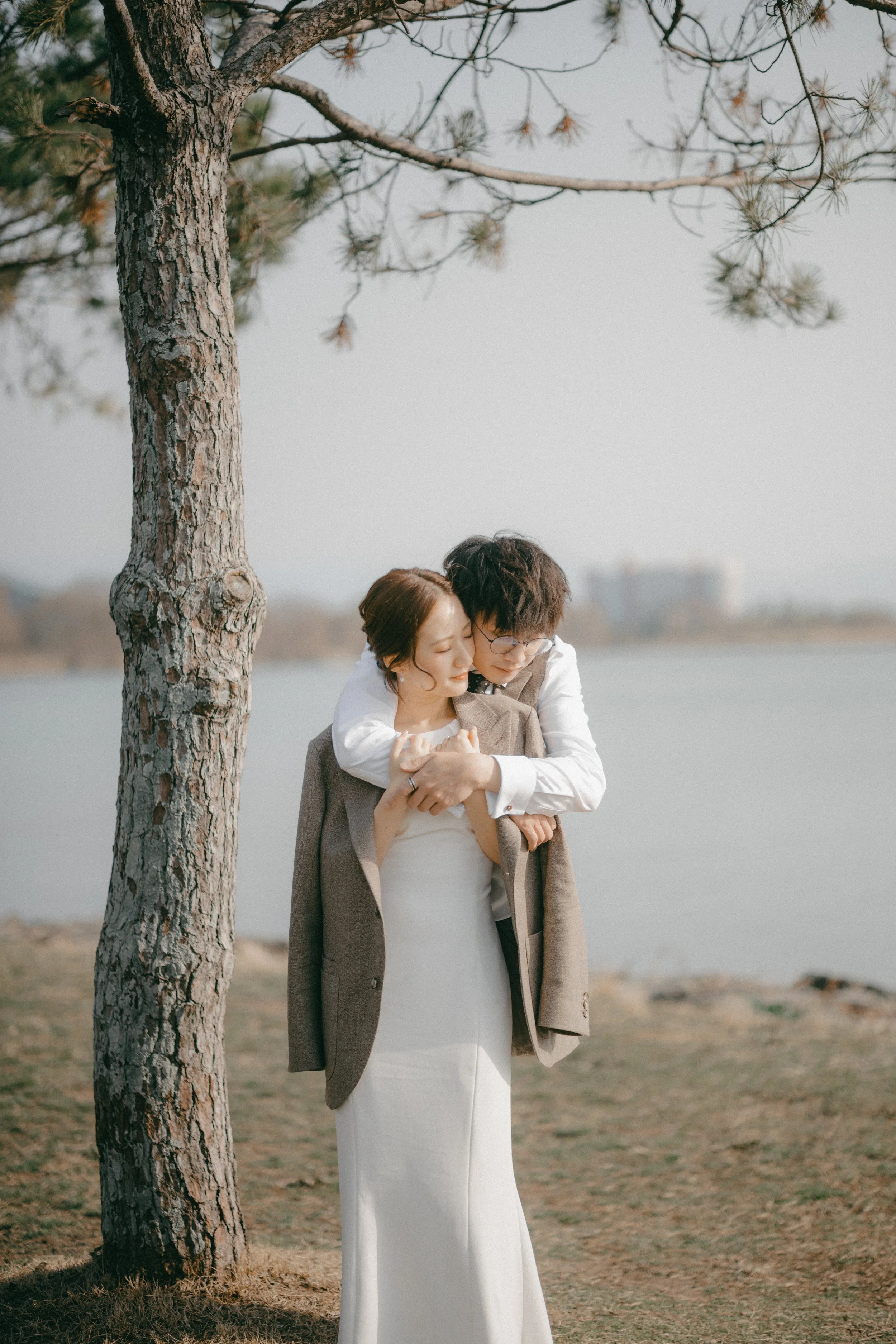 A woman in a white dress and a brown blazer with an overcoat, holding hands, stands under a pine tree by a lake, while a man in glasses, a white shirt, and gray vest embraces her from behind, resting his head on her shoulder. They are outdoors on a c