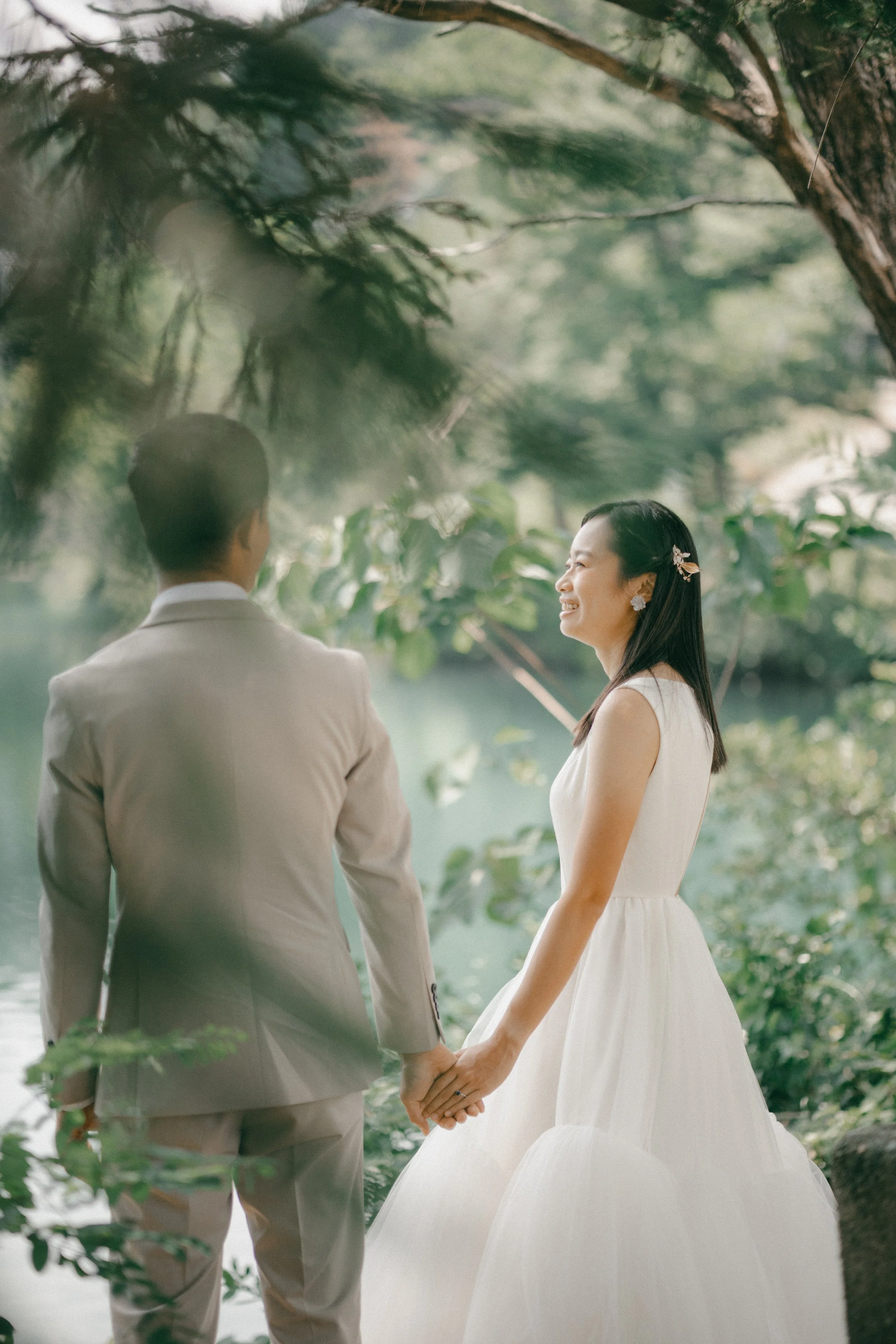 A bride and groom holding hands outdoors near a lake, surrounded by trees, with trees hanging over them.
