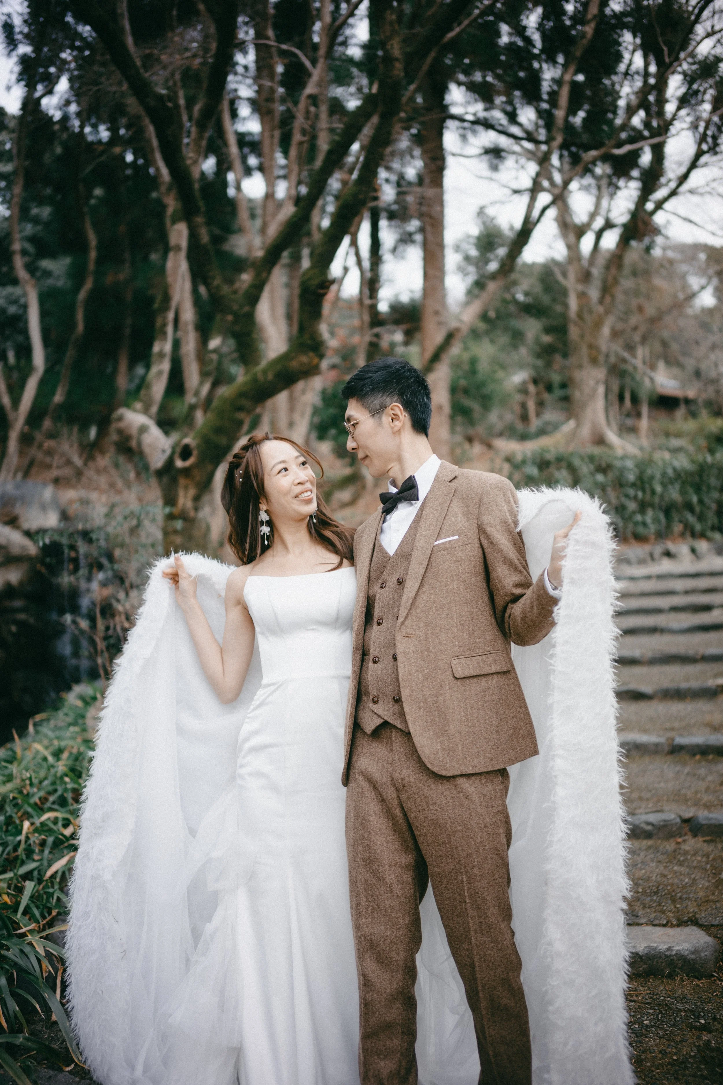 A couple dressed in wedding attire standing outdoors, with trees and a staircase in the background.