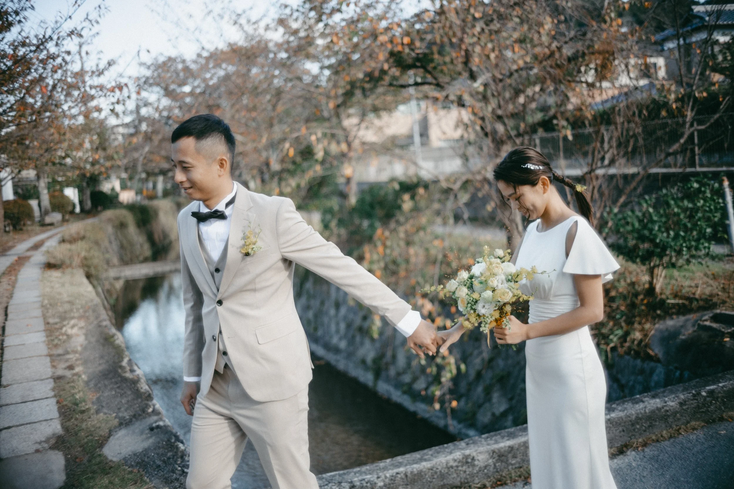 A bride and groom holding hands outdoors near a small stream, with trees and autumn foliage in the background.