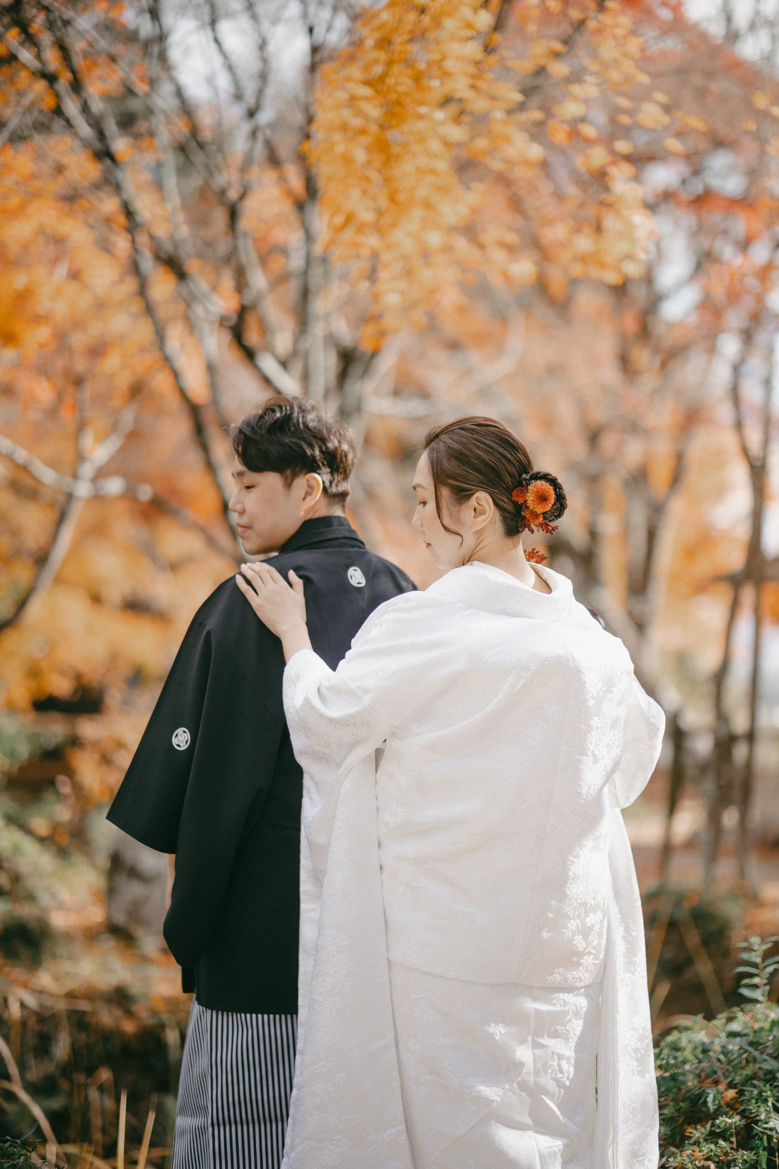 A woman in a traditional white kimono gently touching a man in a black kimono outdoors during autumn, surrounded by orange and yellow fall foliage.