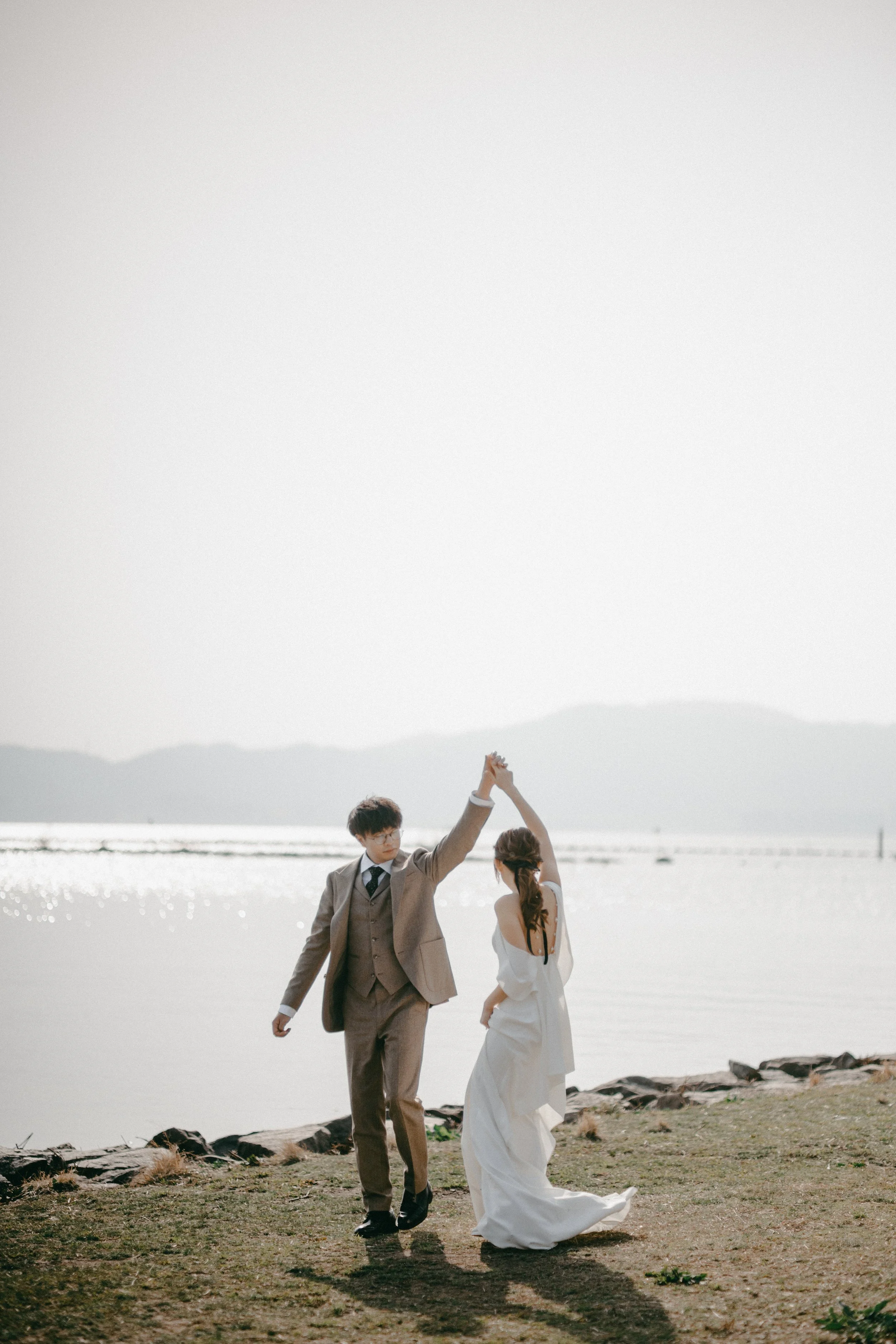 A couple dressed in wedding attire dancing by a lakeshore with mountains in the background.