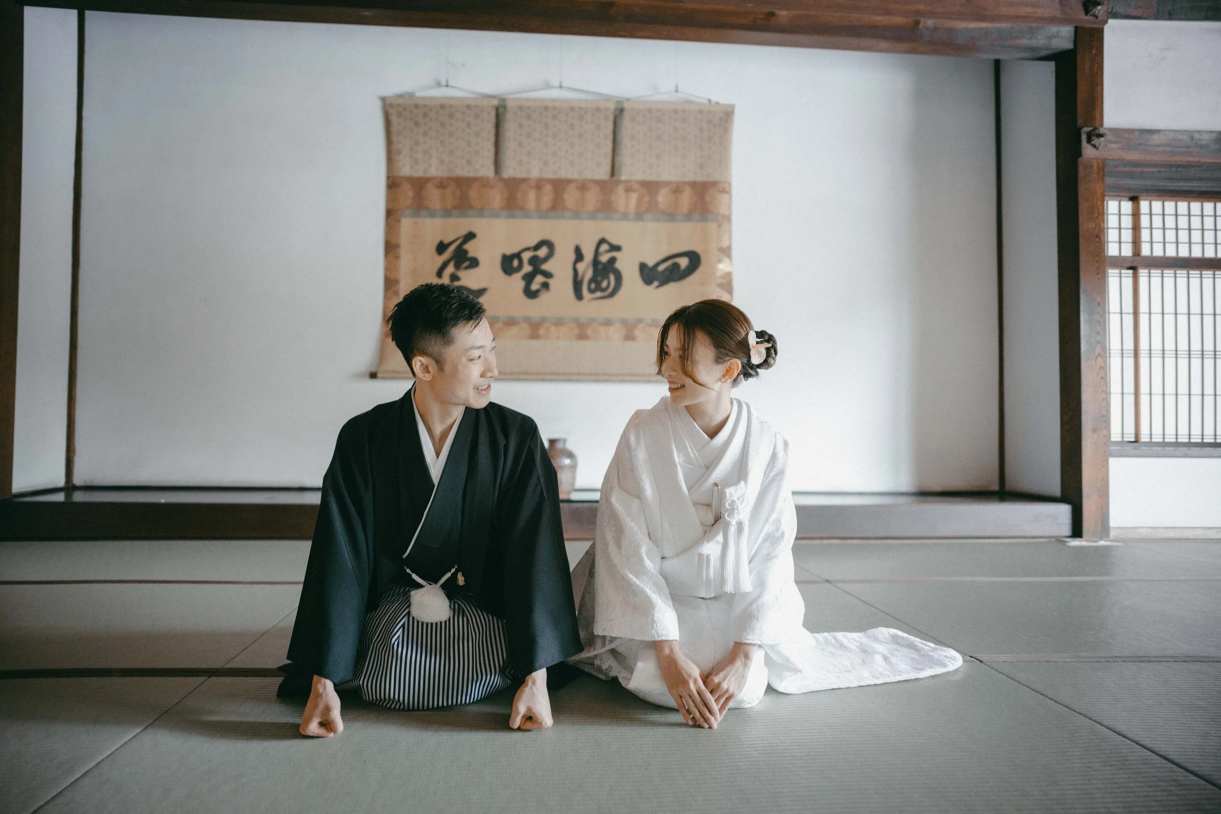 A man and woman dressed in traditional Japanese kimonos sitting on a tatami mat in a Japanese-style room, smiling at each other.