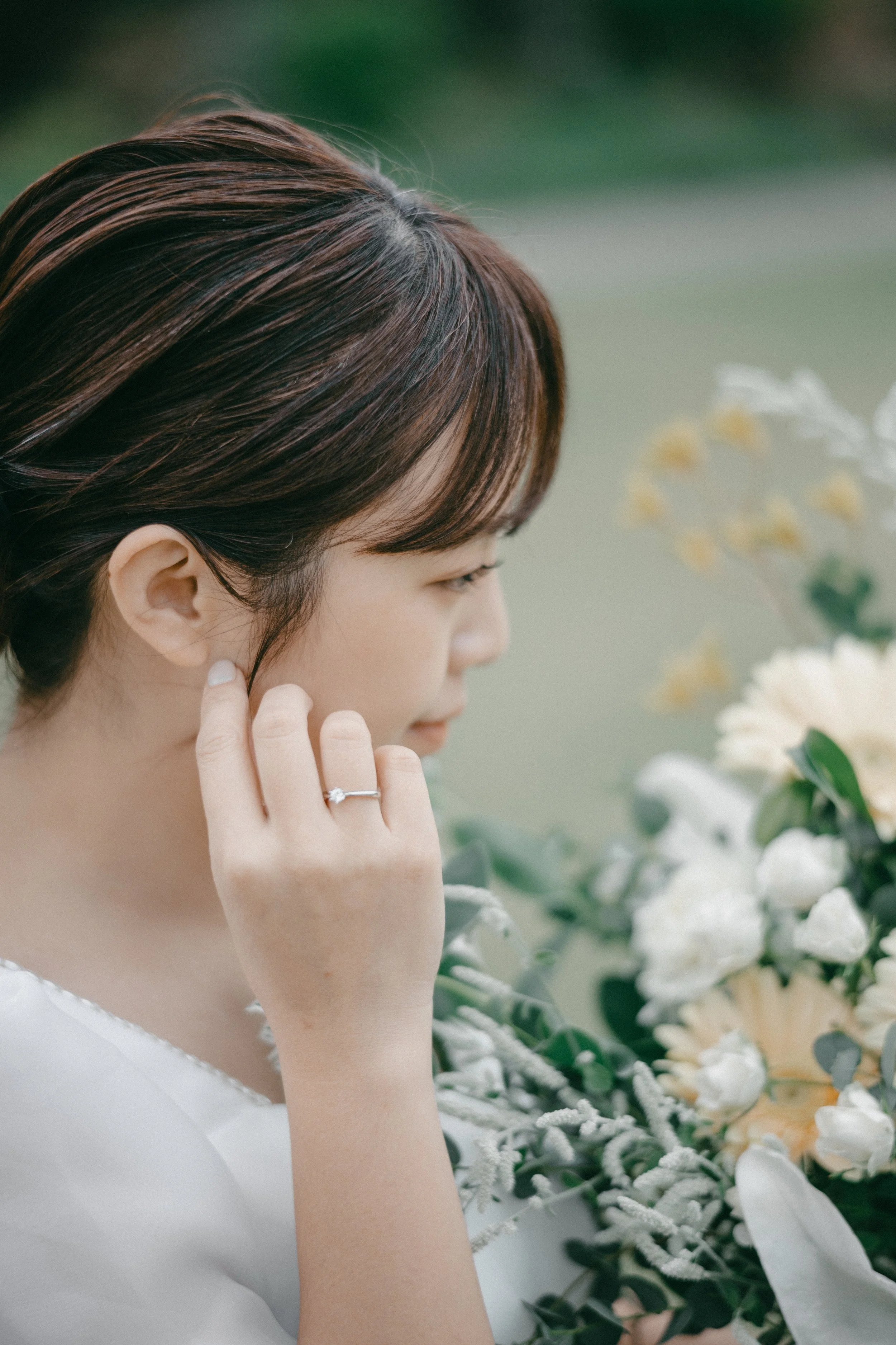 A woman with short brown hair and a ring on her finger touches her face, standing near a bouquet of flowers outdoors.