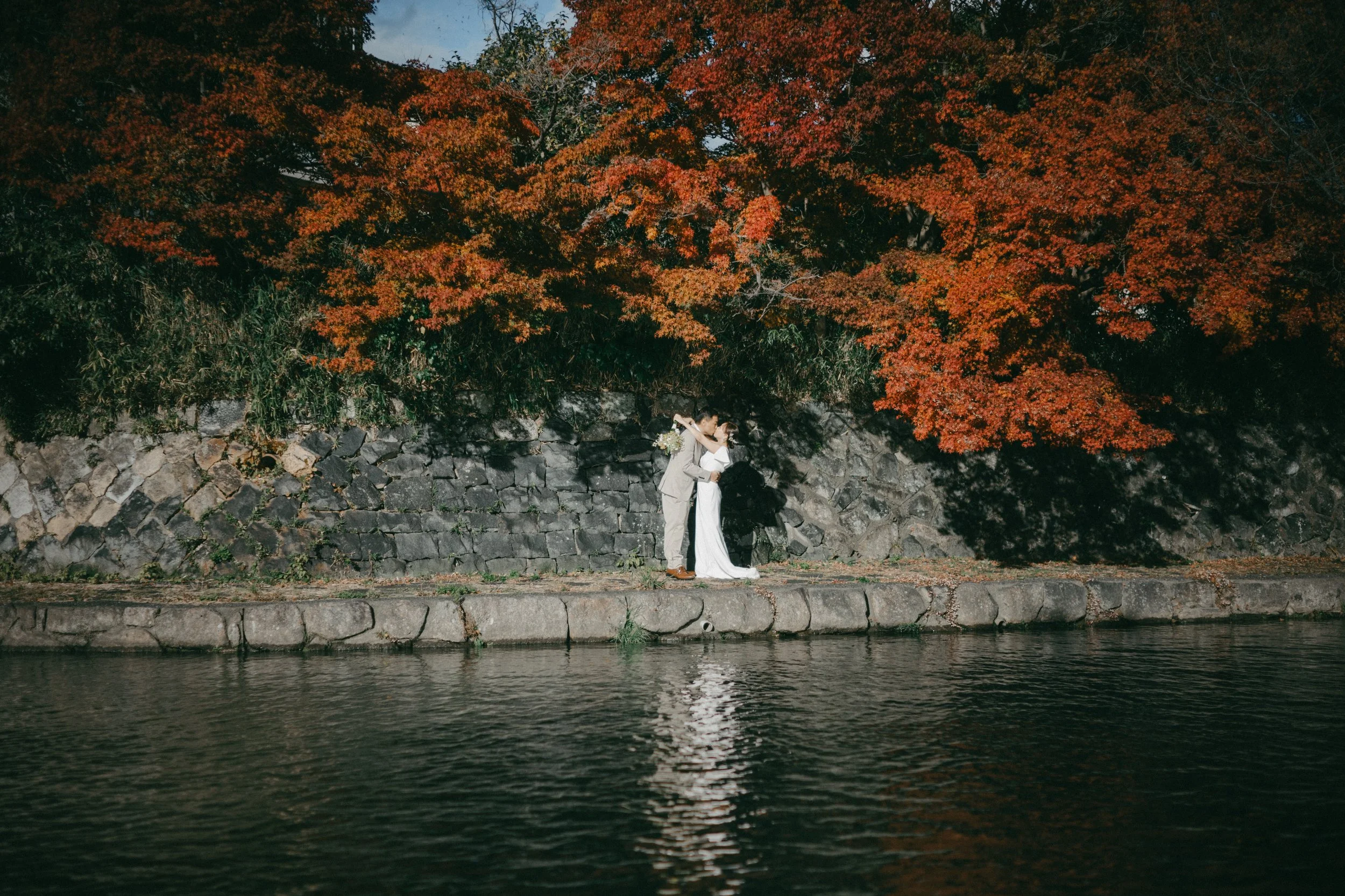 A couple in wedding attire standing on a stone path beside a body of water, embracing and kissing, with large orange and red fall foliage trees behind them.
