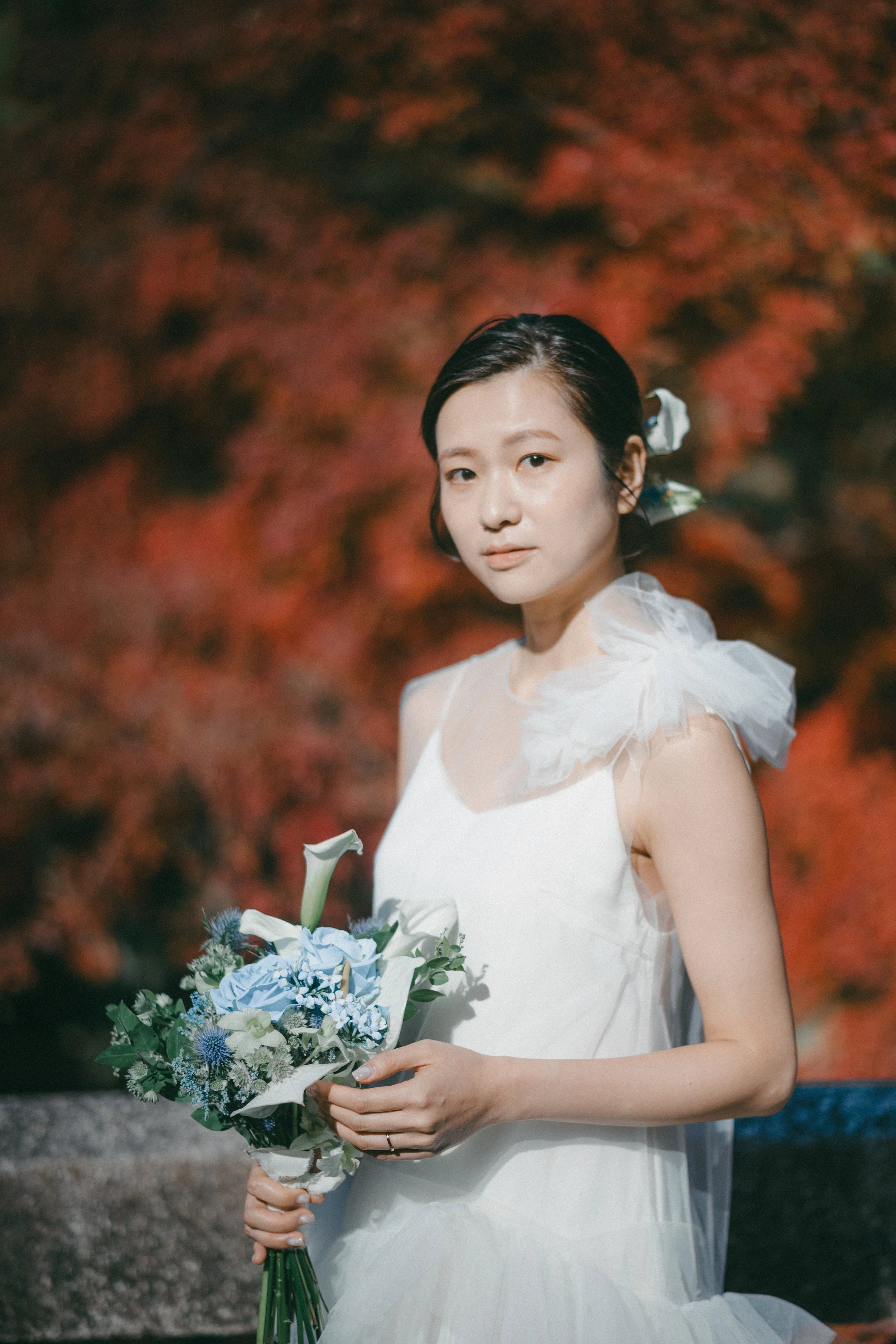 A woman in a white dress holding a bouquet of white and blue flowers, standing in front of a blurred red and orange foliage background.