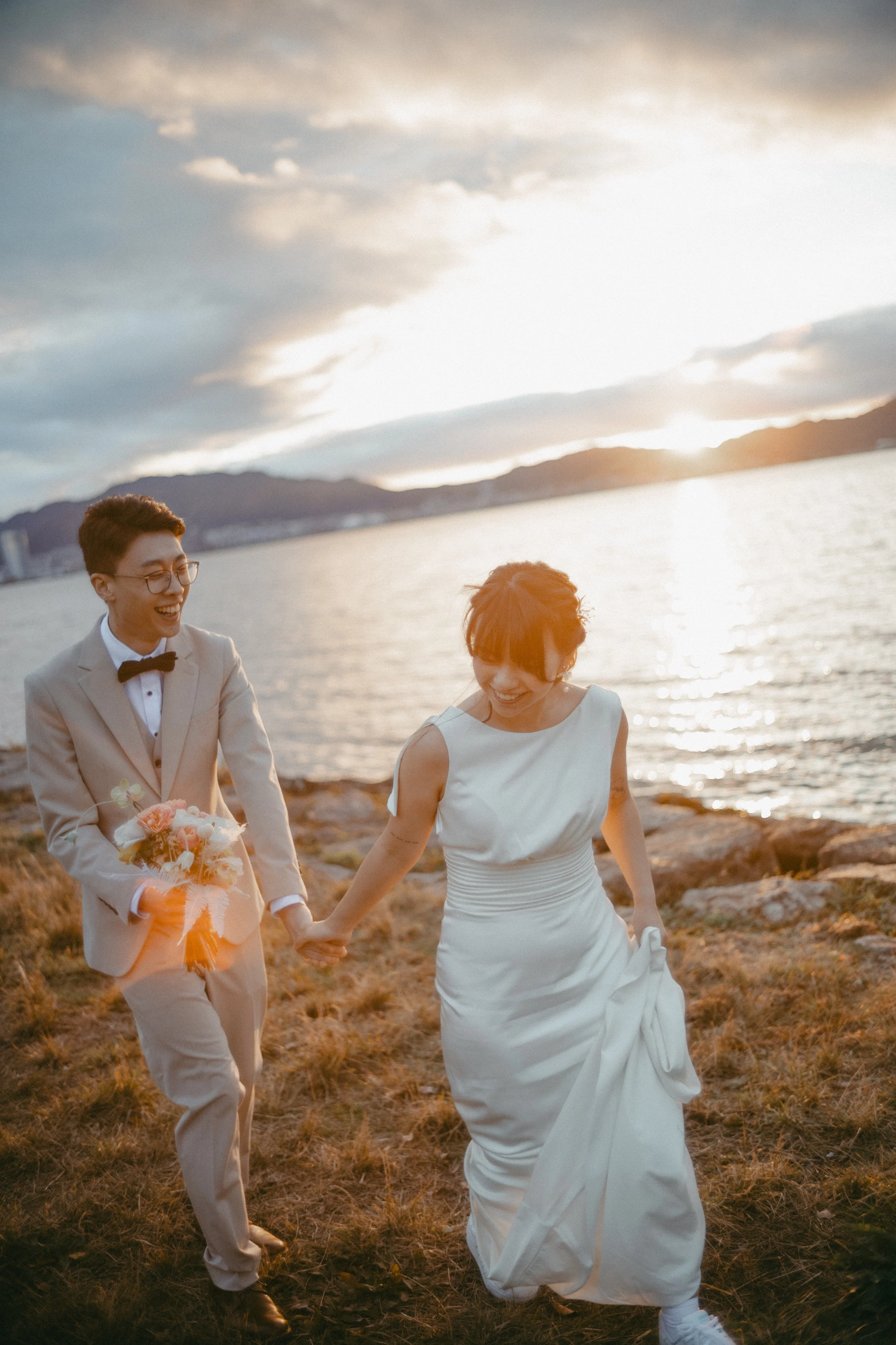 A joyful couple in wedding attire holding hands and laughing near a body of water at sunset.