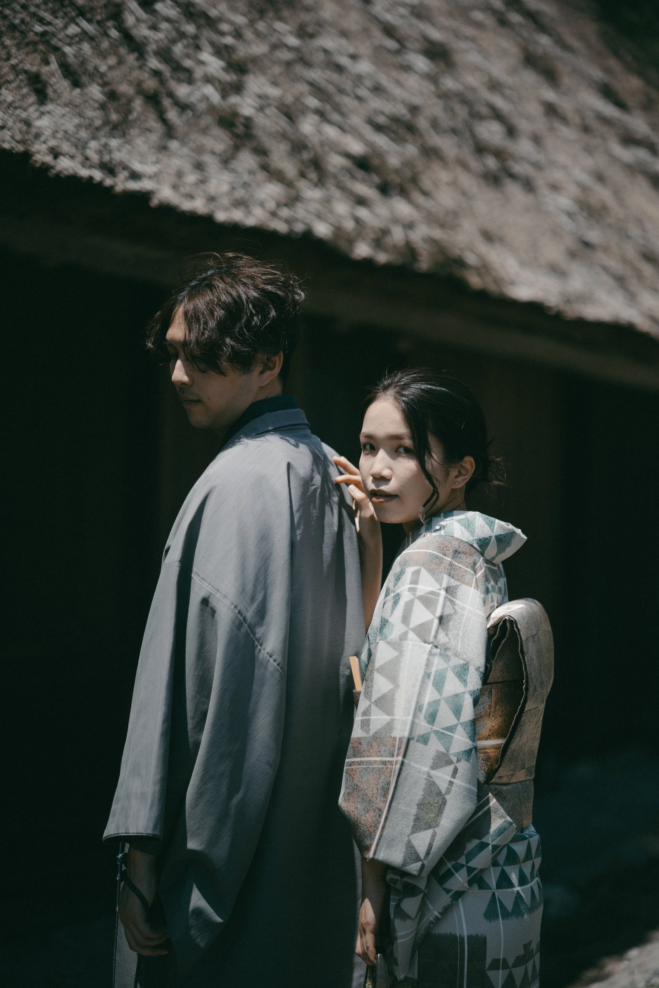 A man and a woman wearing traditional Japanese kimonos stand together in a dark, cave-like setting. The woman looks at the camera, while the man looks downward.