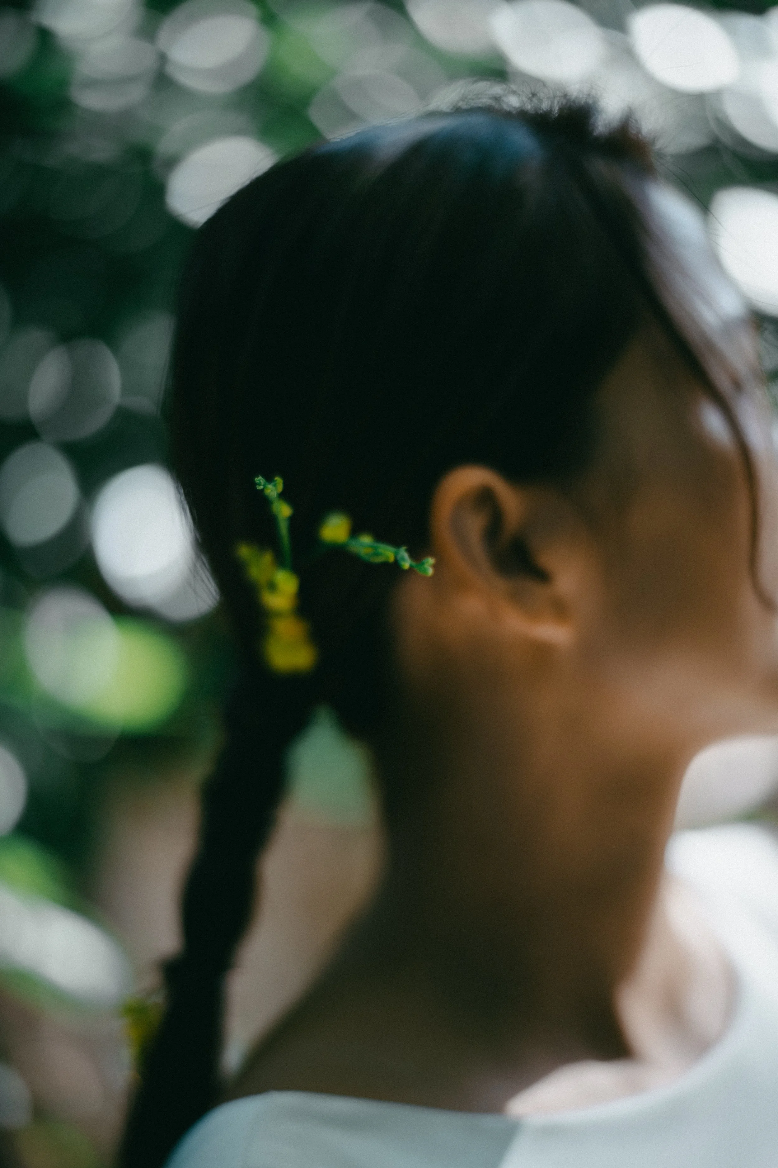 A woman with dark hair styled in a braid, wearing a small green decorative hair accessory, with a blurred background of green and white bokeh lights.