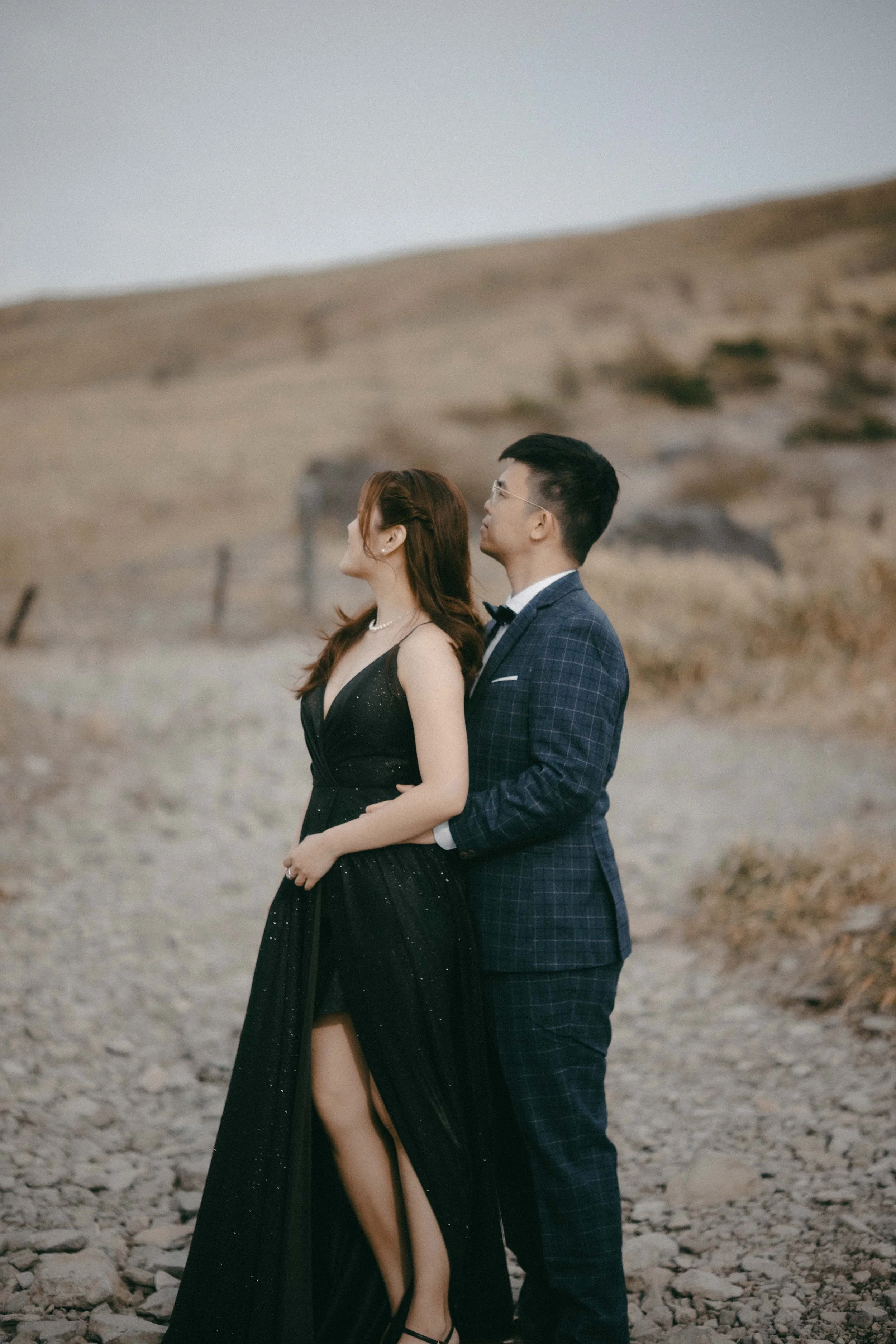 A couple standing close together outdoors in a rocky, hilly landscape, dressed in formal attire, with the woman wearing a black gown and the man in a checkered suit, facing away from the camera.