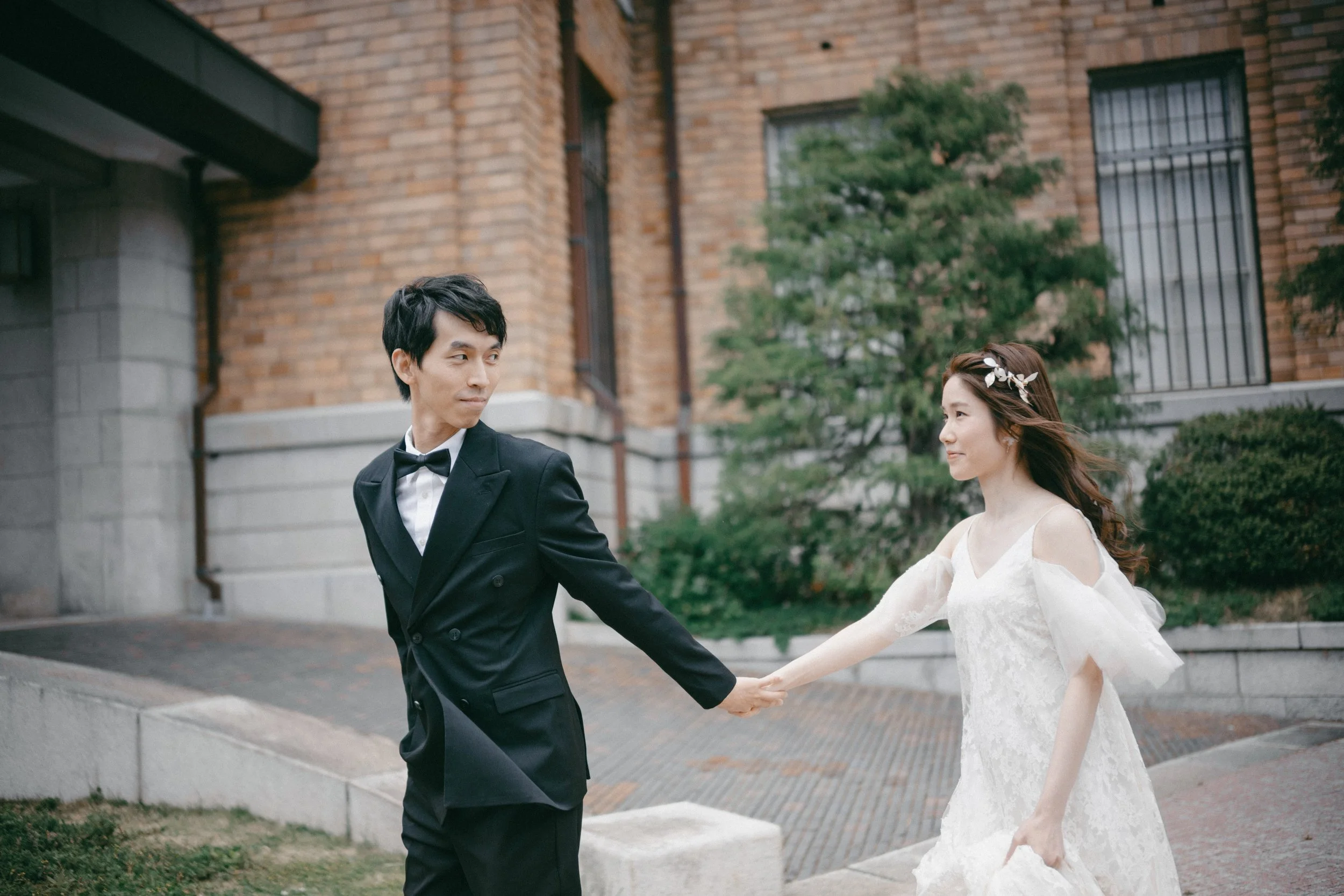 A bride and groom holding hands outdoors with a brick building and greenery in the background.