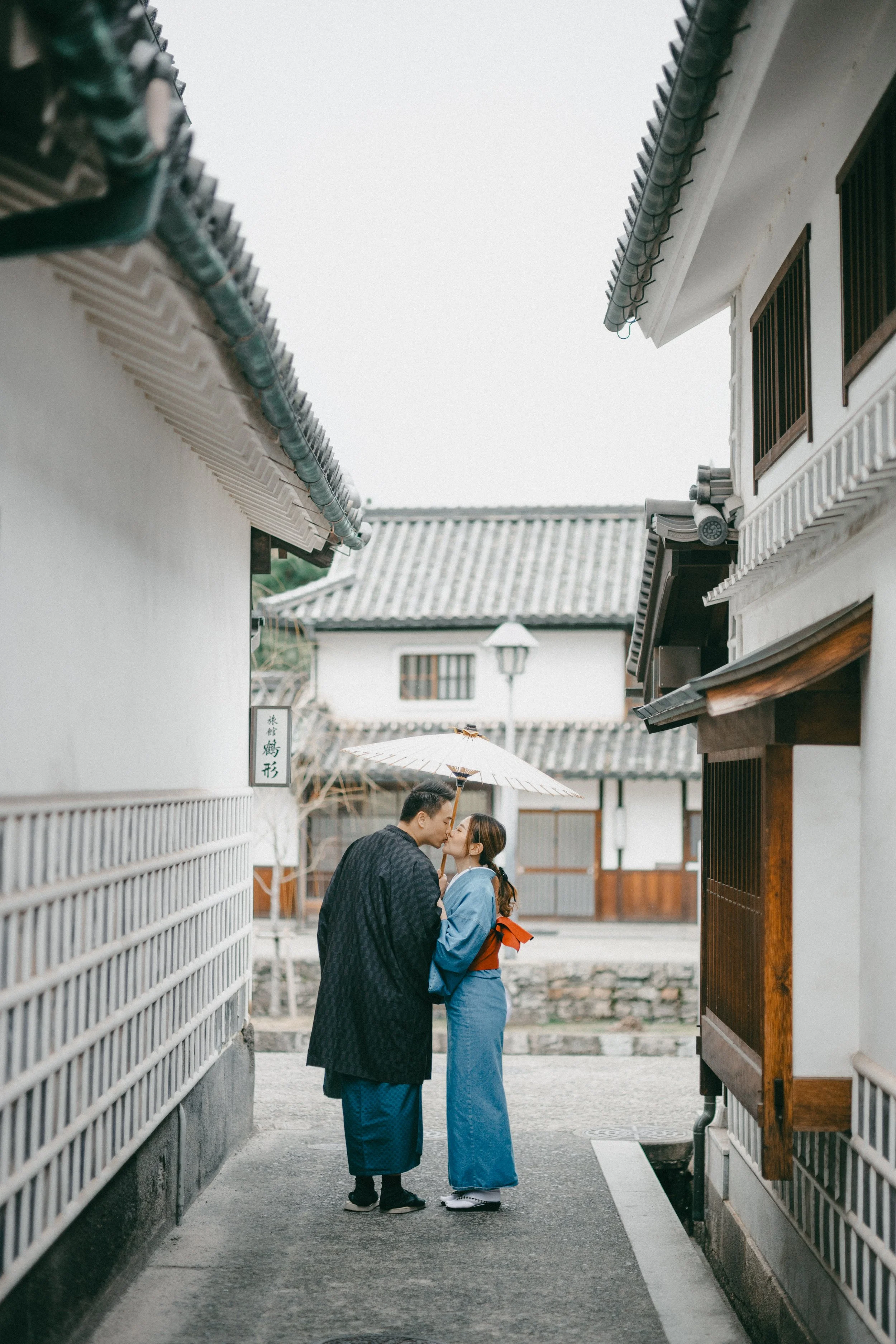 A couple dressed in traditional Japanese attire sharing a kiss under an umbrella in a narrow alley with white buildings and tiled roofs.