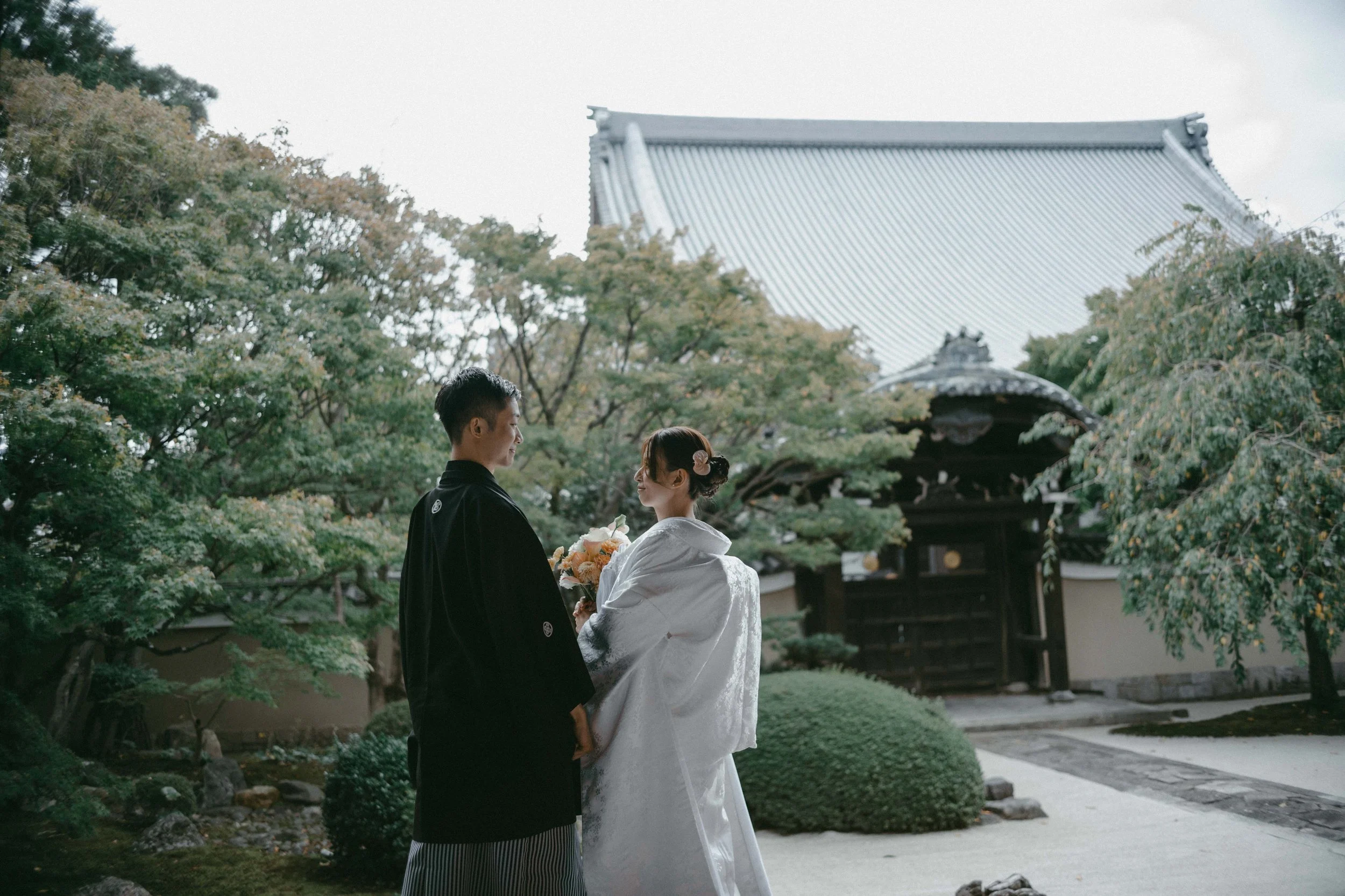 A couple in traditional Japanese wedding attire stands in a garden with lush greenery, trees, and a traditional building with a tiled roof in the background.