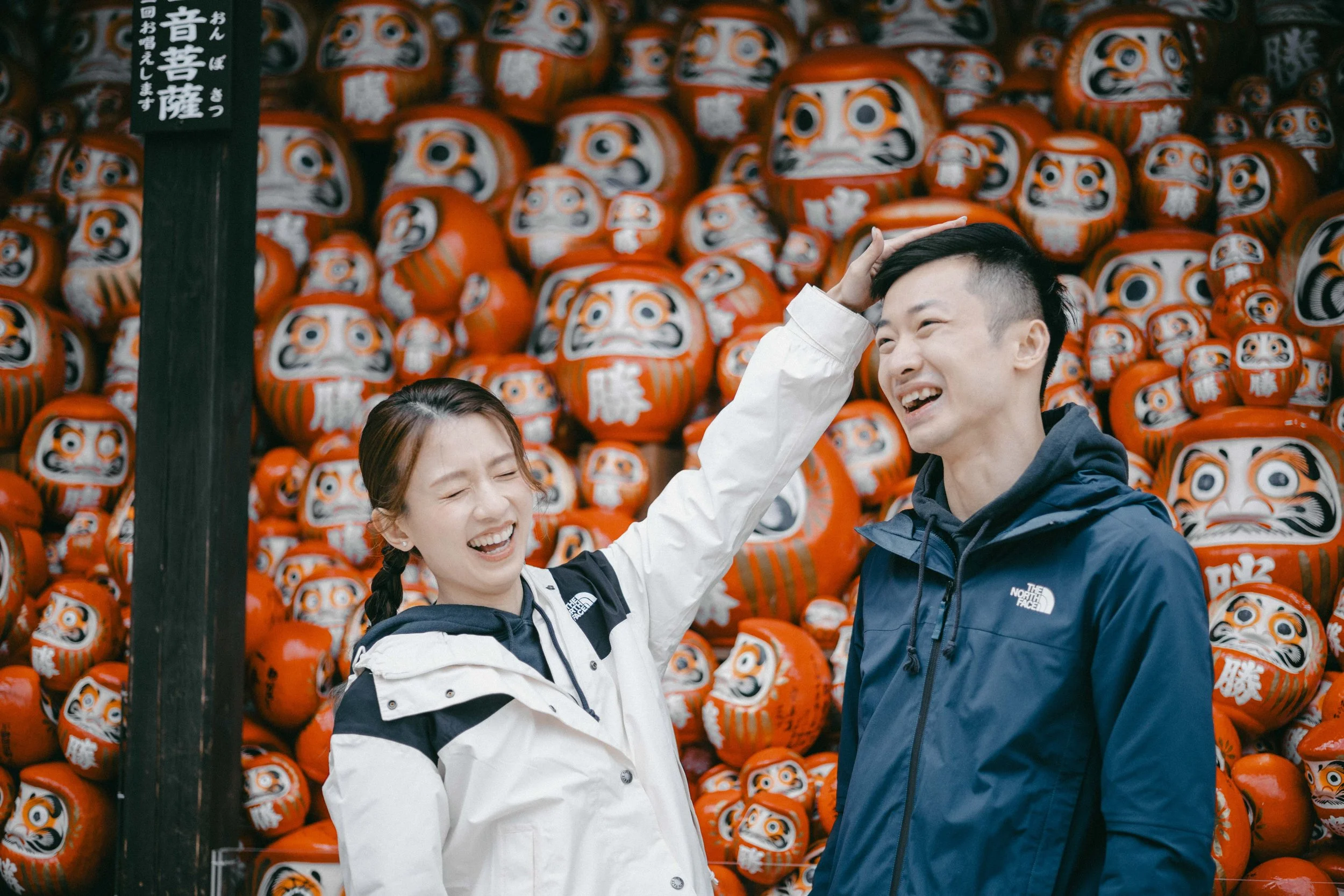 A woman and a man are smiling and laughing in front of a wall of red Daruma dolls. The woman is touching the man's head.