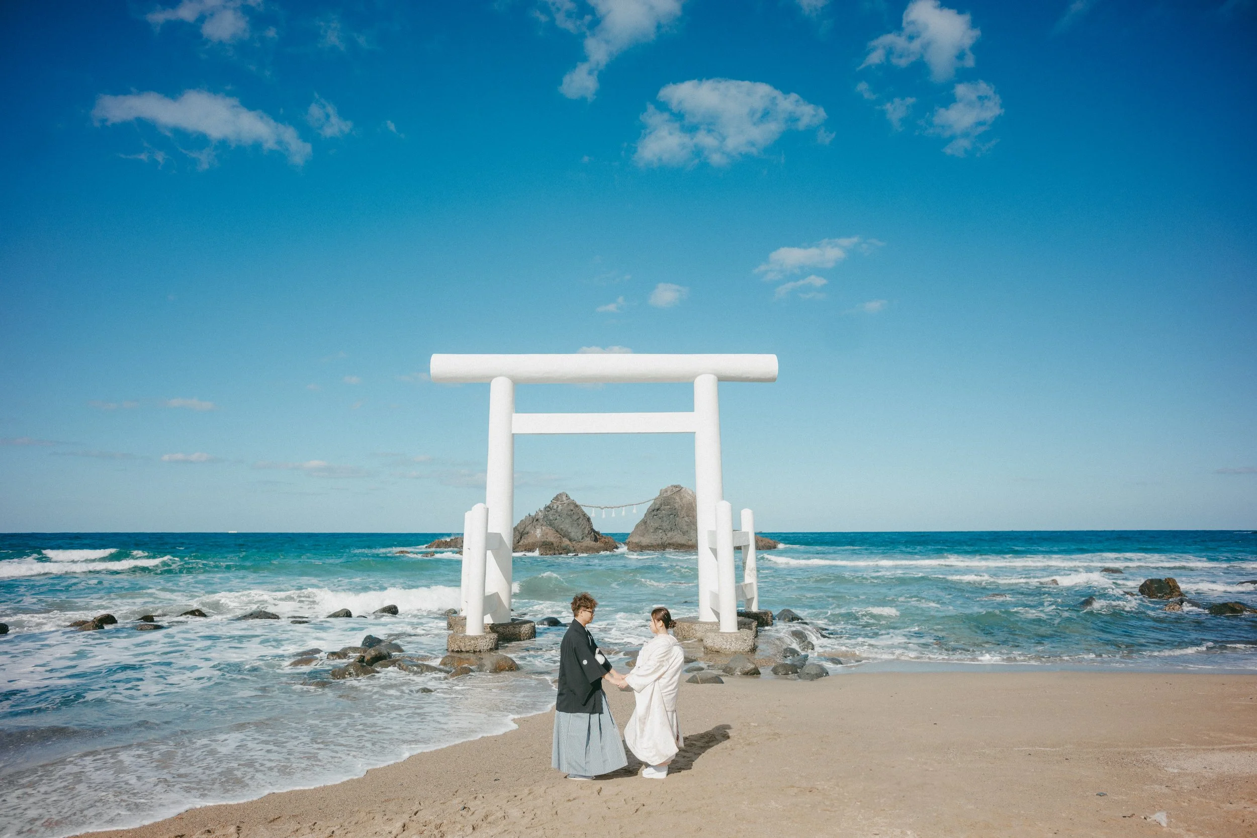 A couple in traditional Japanese attire holding hands on a beach with a large white torii gate in the background, near the ocean and rocks under a blue sky with clouds.