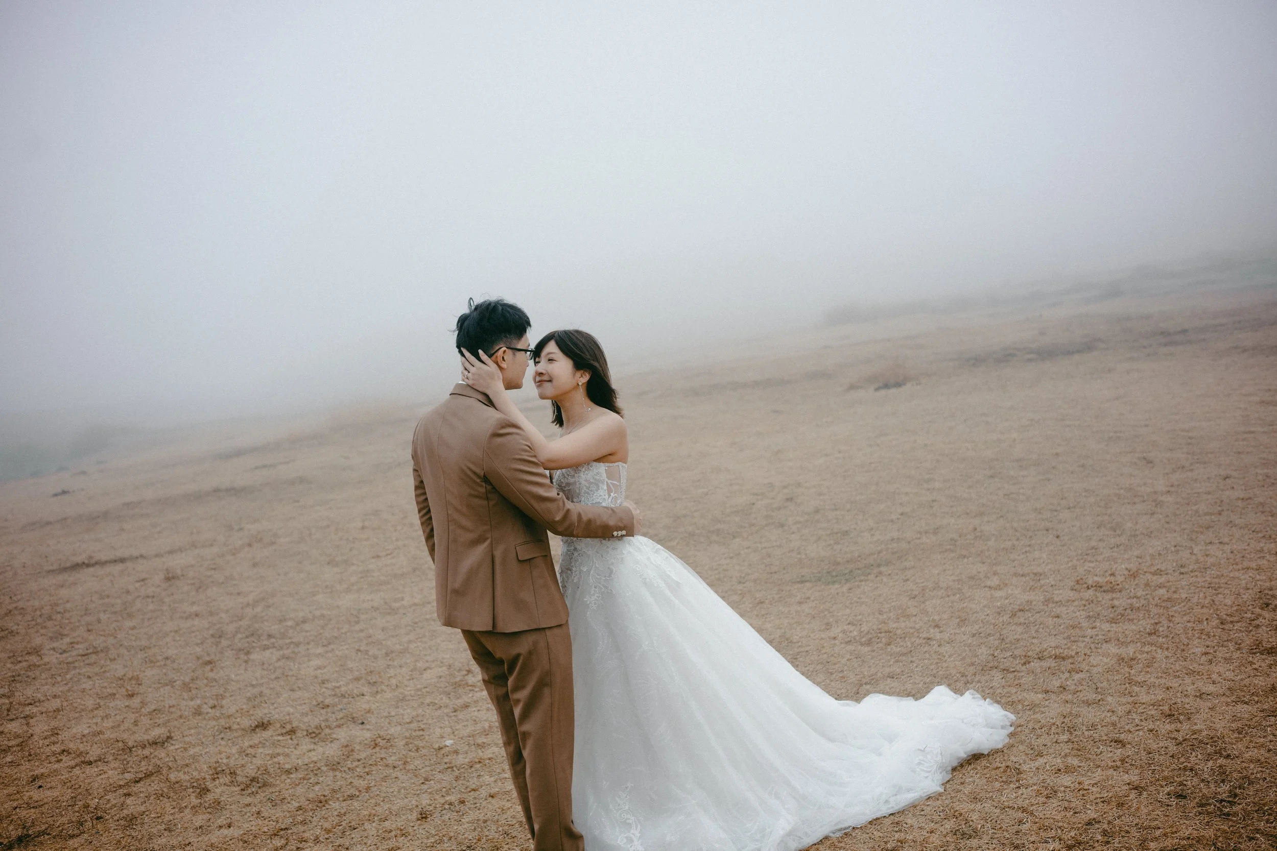 A bride and groom in wedding attire standing close together on a foggy, barren landscape with brown ground, smiling and touching faces.
