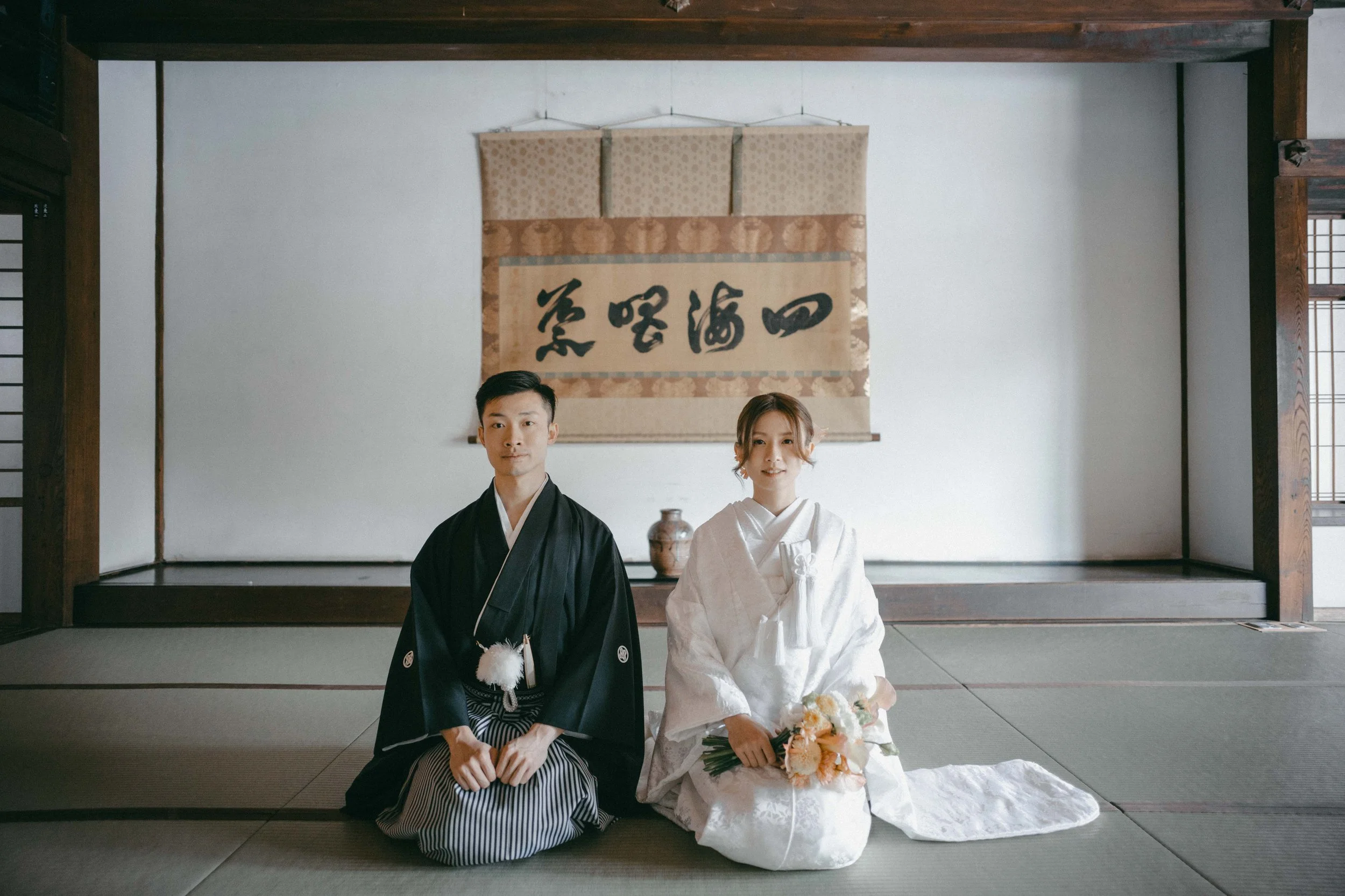 A couple dressed in traditional Japanese wedding attire sitting on tatami mats in a room with minimalist decor, a calligraphy scroll hanging on the wall behind them, and a vase on a raised platform.