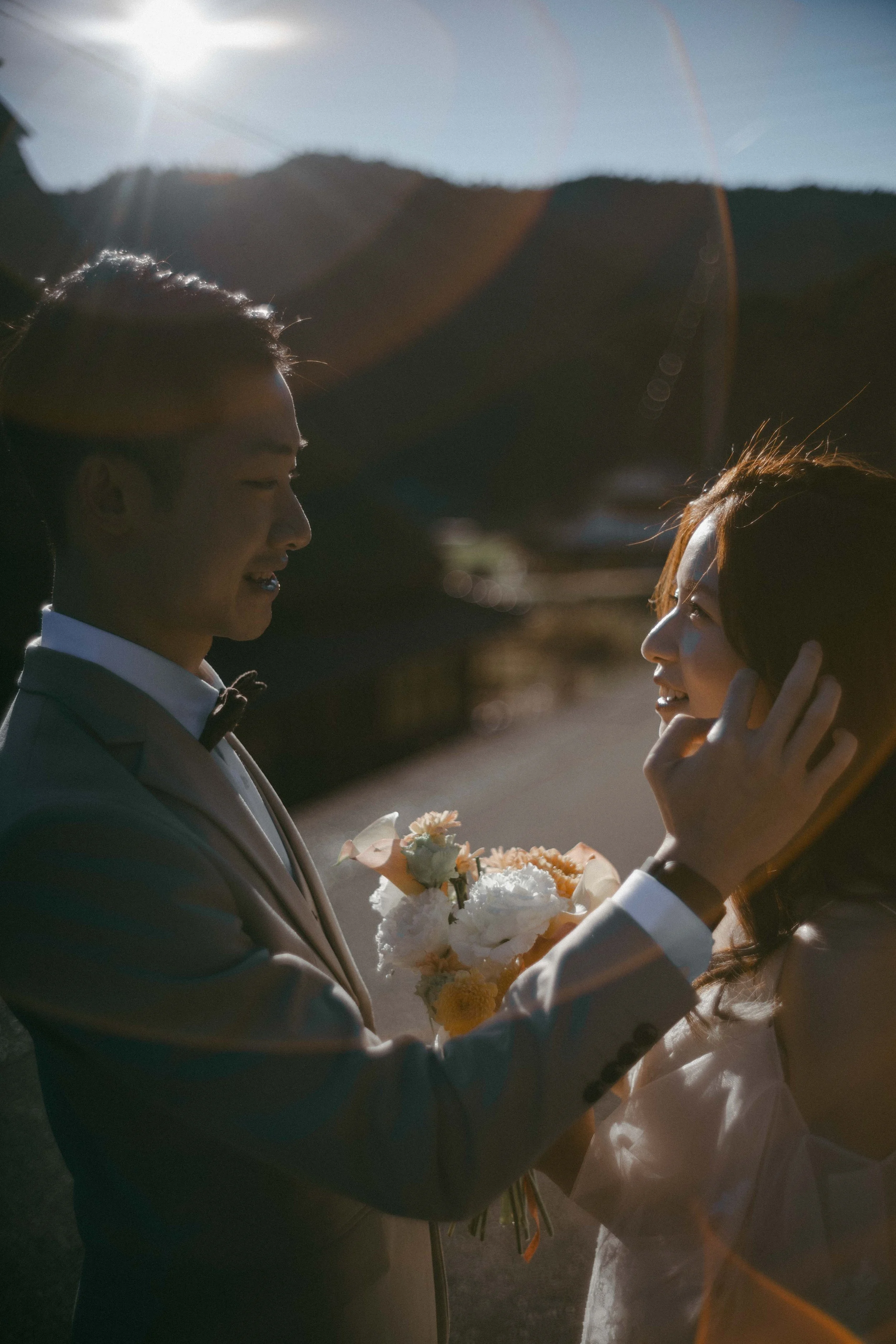 A couple in wedding attire standing outdoors during sunset, with the groom gently holding the bride's face and holding a bouquet of flowers.