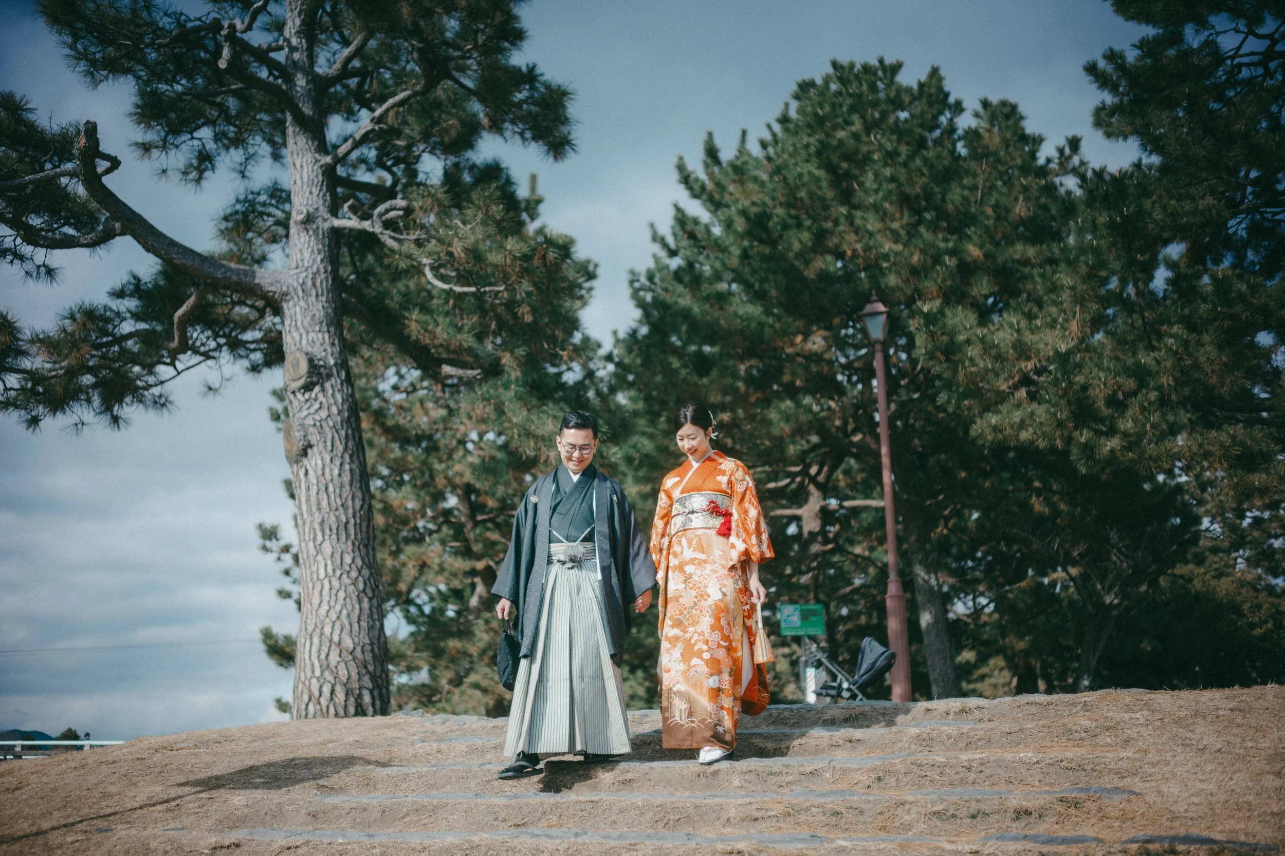 A couple dressed in traditional Japanese wedding attire walking down stone steps outdoors surrounded by tall trees.