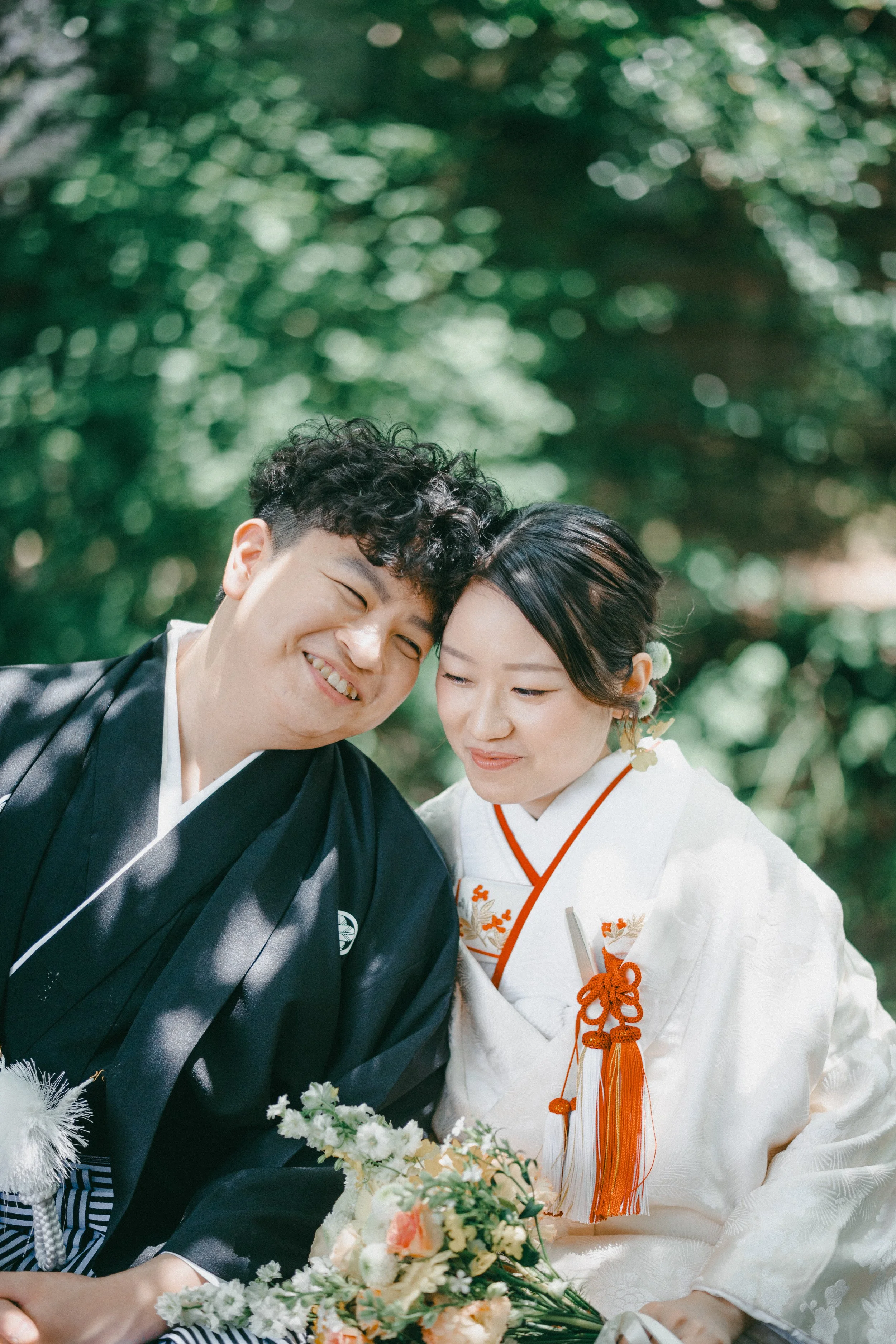 A smiling man and woman in traditional Japanese wedding attire, sitting closely outdoors with green foliage in the background, holding a bouquet of flowers.