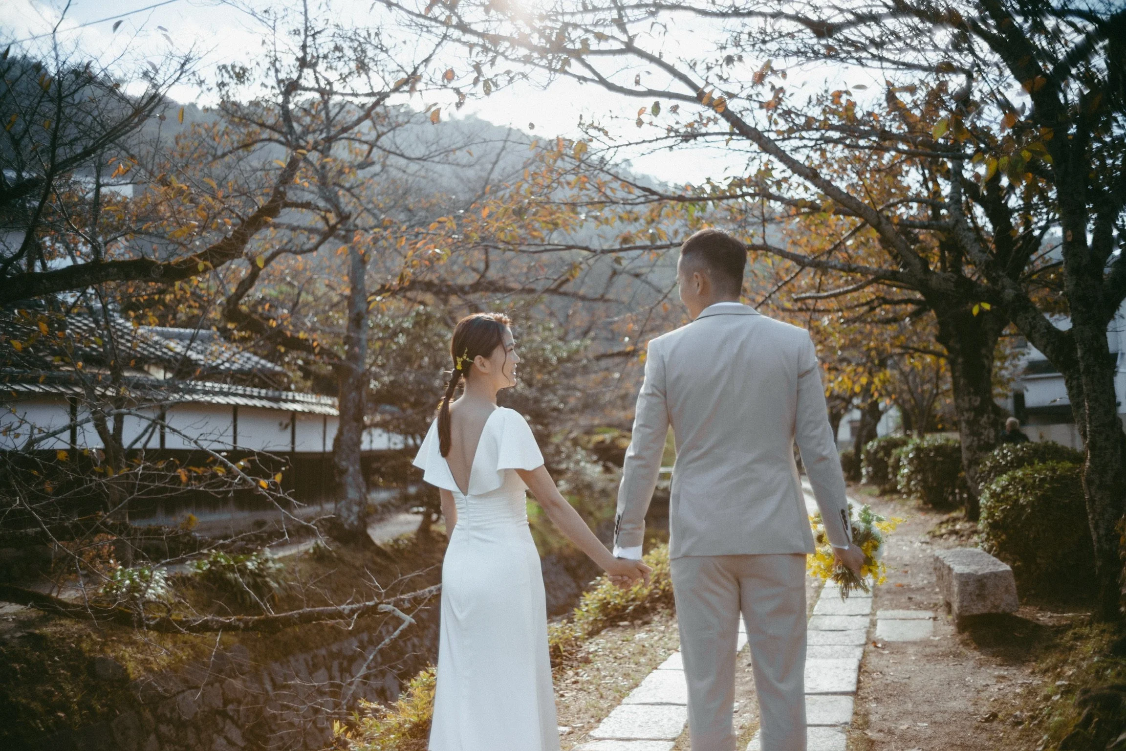 A couple dressed in wedding attire holding hands and walking outdoors on a stone pathway through a park with leafless trees and a traditional building in the background.