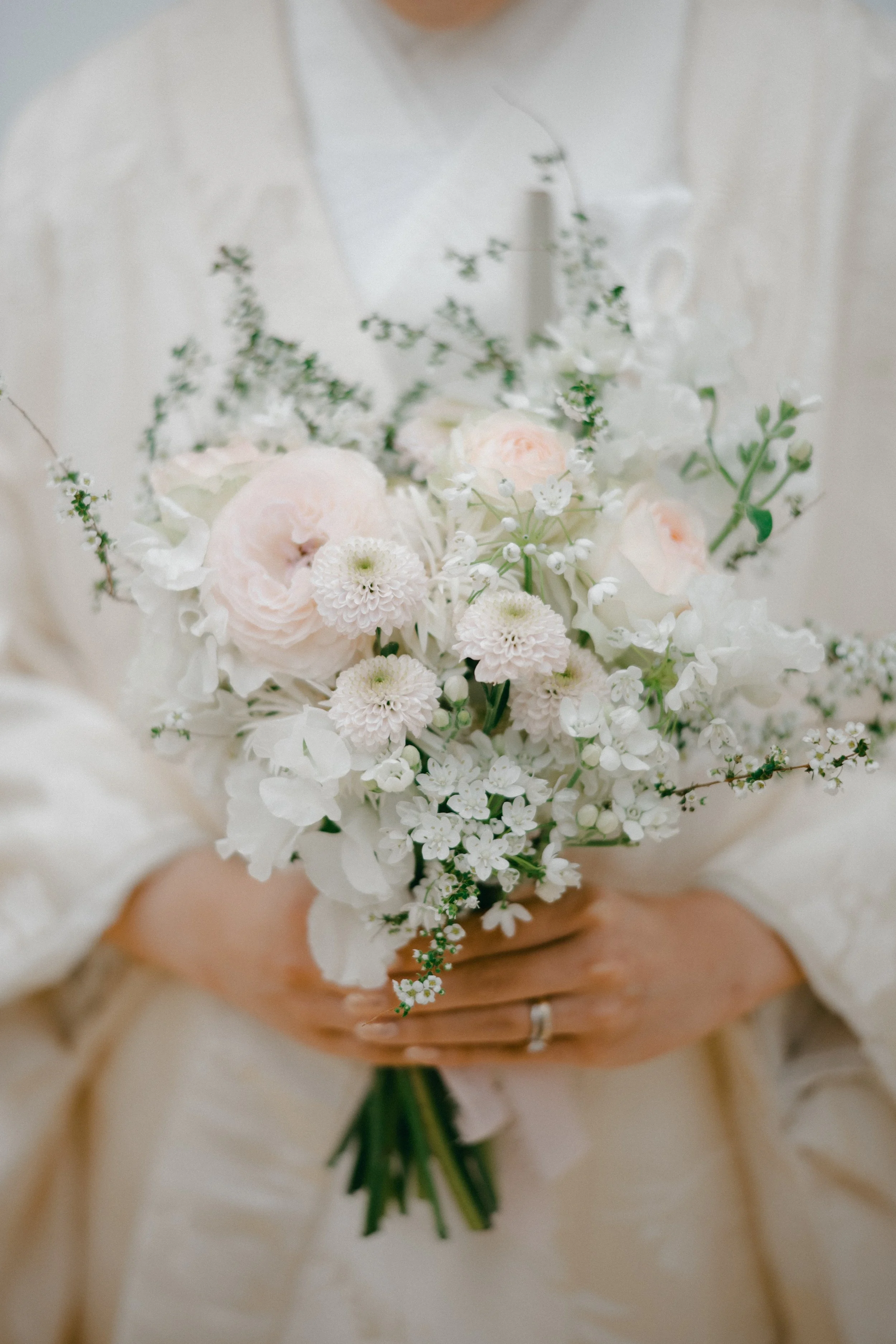 Person holding a bouquet of white and light pink flowers, including roses, chrysanthemums, and small blossoms, in a cream-colored dress.