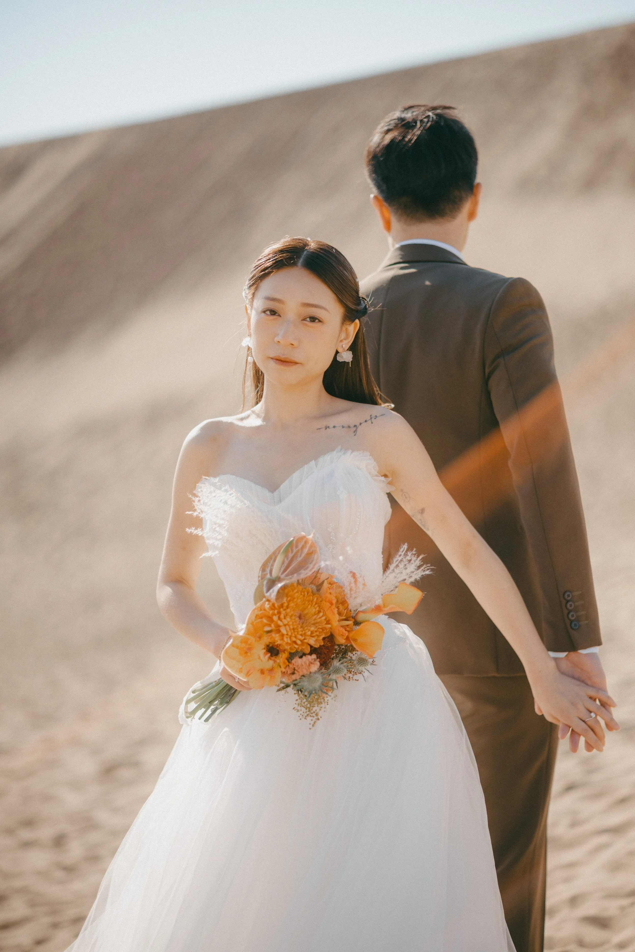 A bride holding a bouquet and standing in front of a groom, on a sandy outdoor setting during daytime.