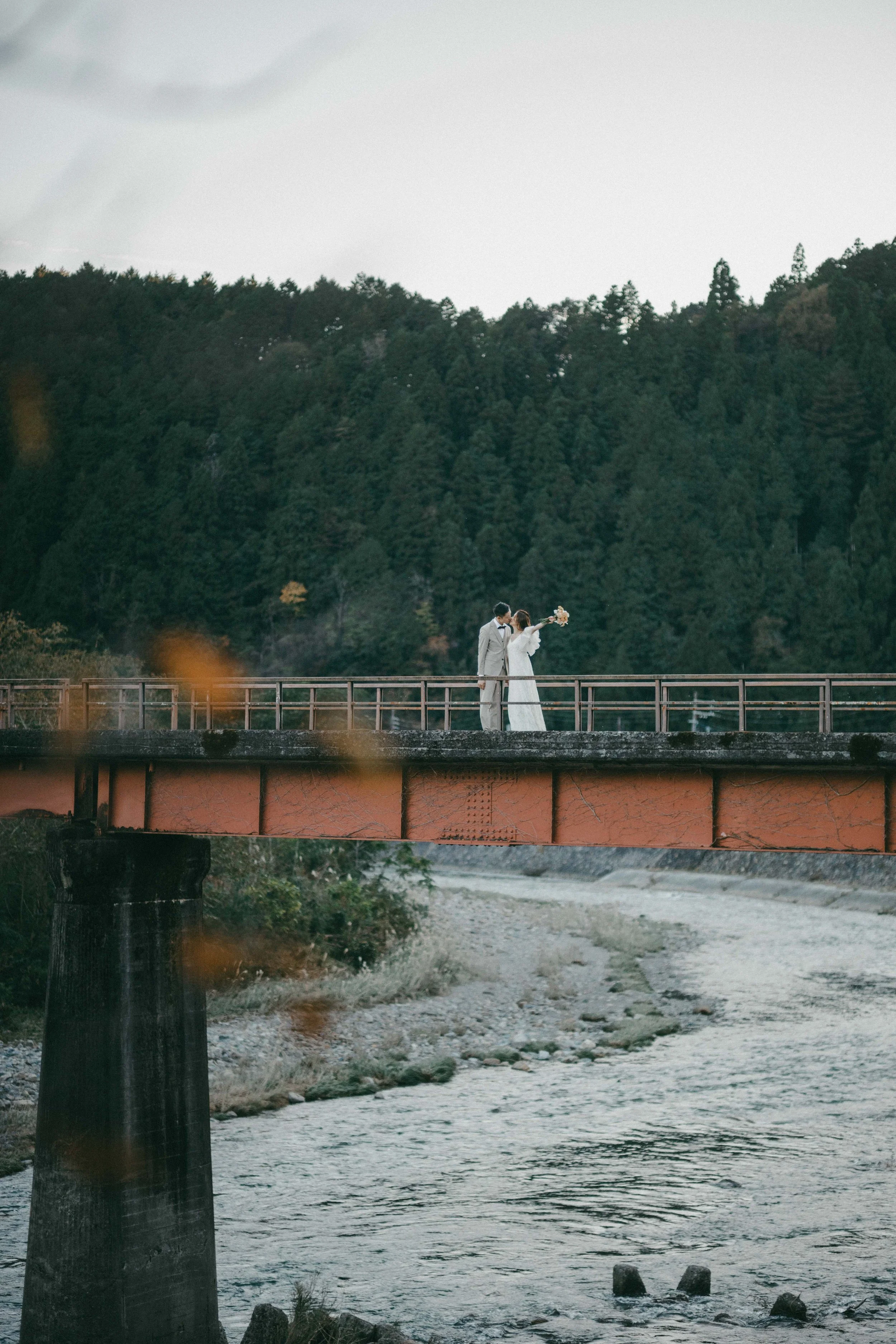 A bride and groom standing on a bridge over a river, with the bride holding a bouquet and the groom dressed in a light-colored suit, against a backdrop of forested hills.