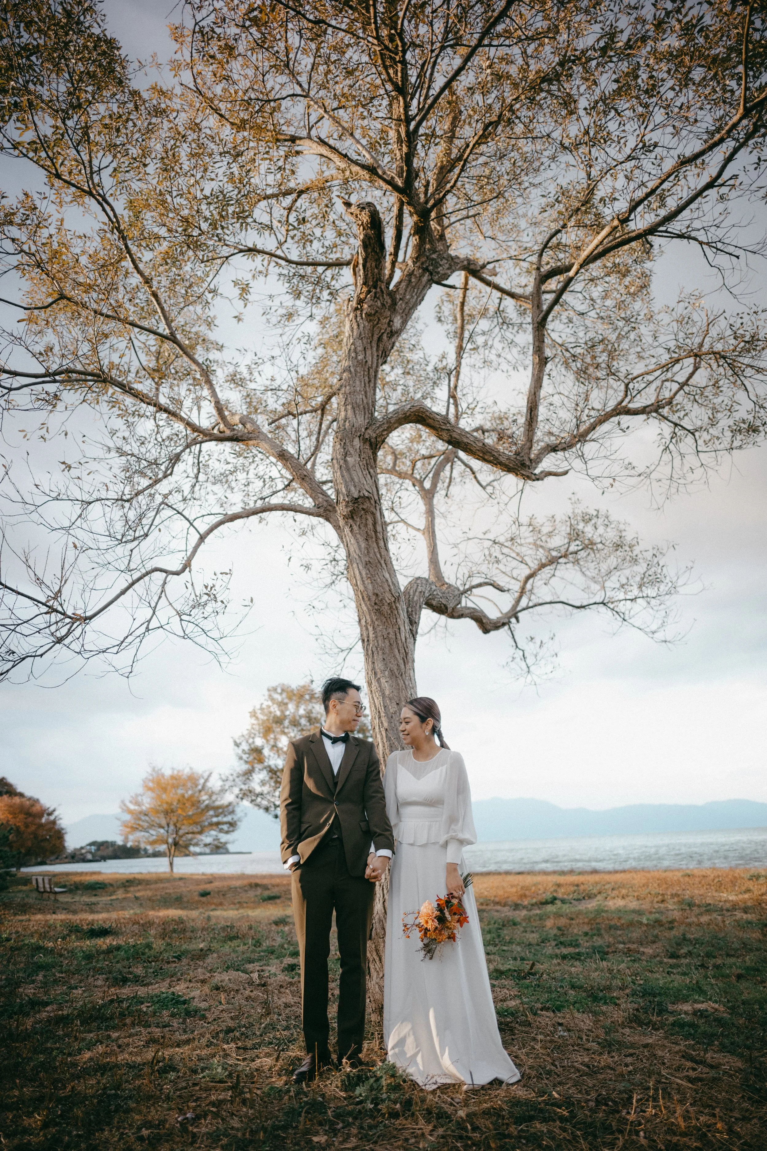 A bride and groom holding hands under a tall tree by a body of water during sunset.
